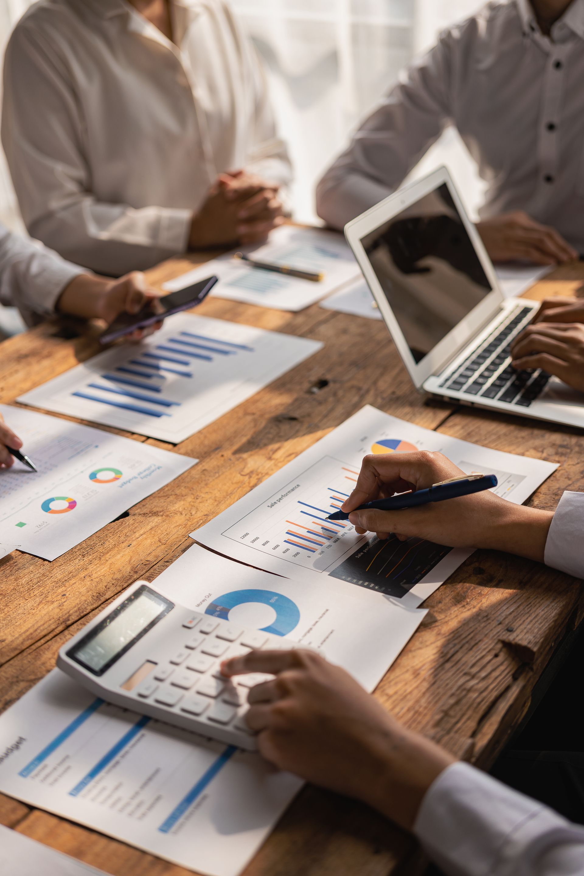 People in a meeting at a wooden table, reviewing data with charts, calculators, and a laptop.