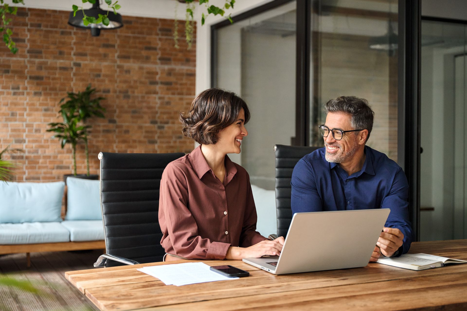Two people at a wooden desk with a laptop, smiling. Brick wall and greenery in the background.
