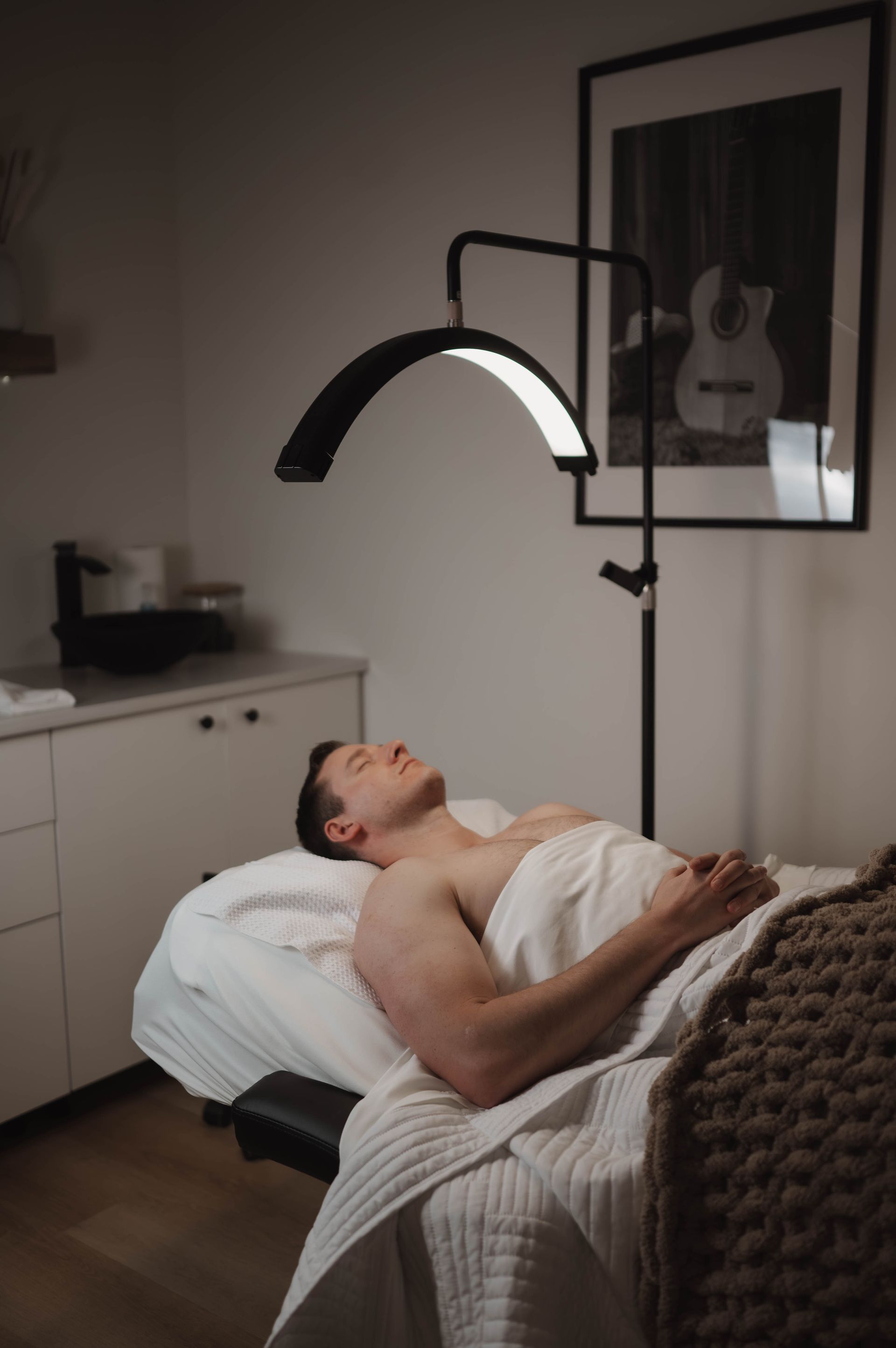 A shirtless man is laying on a massage table under a lamp.