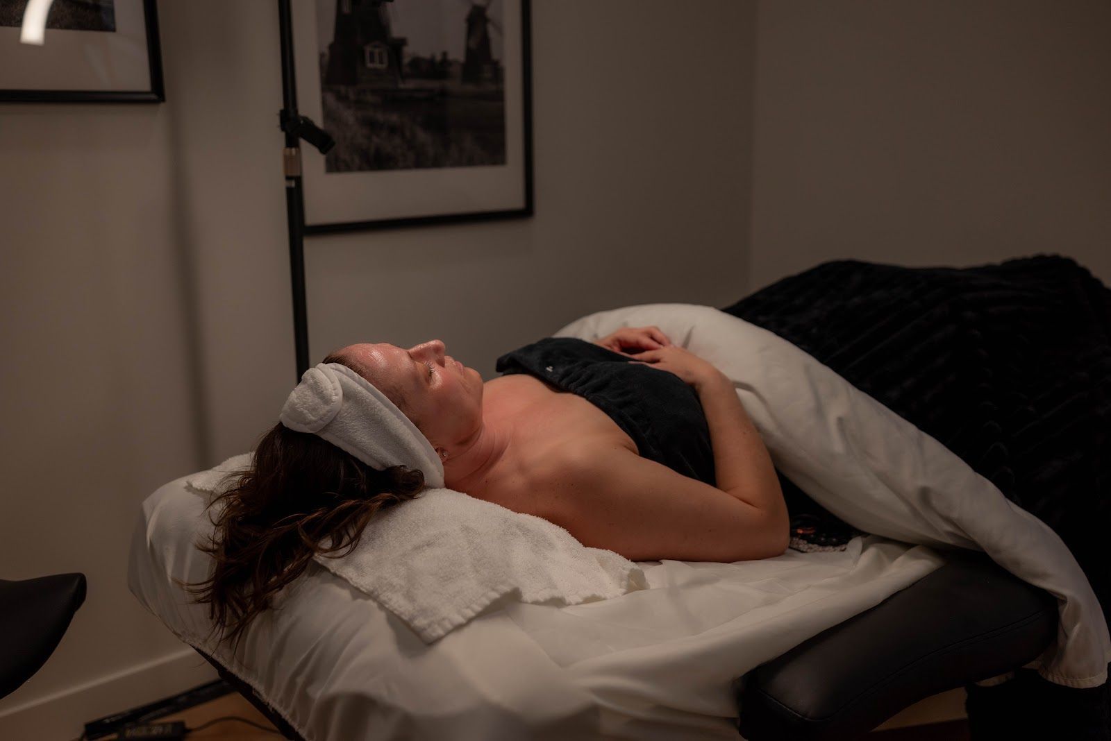 Woman relaxing on a massage table, covered with a blanket. She's wearing a headband in a dimly lit room.