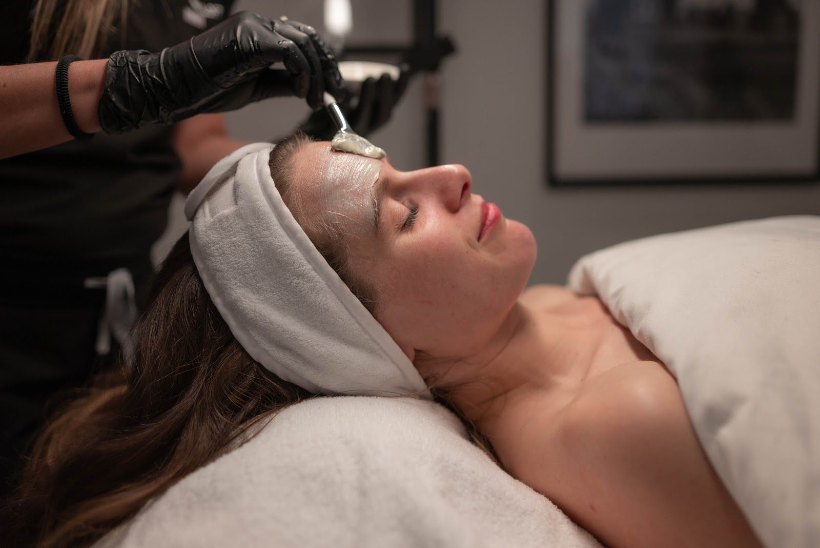 Person getting facial treatment at spa; technician applies product to face with black gloves.