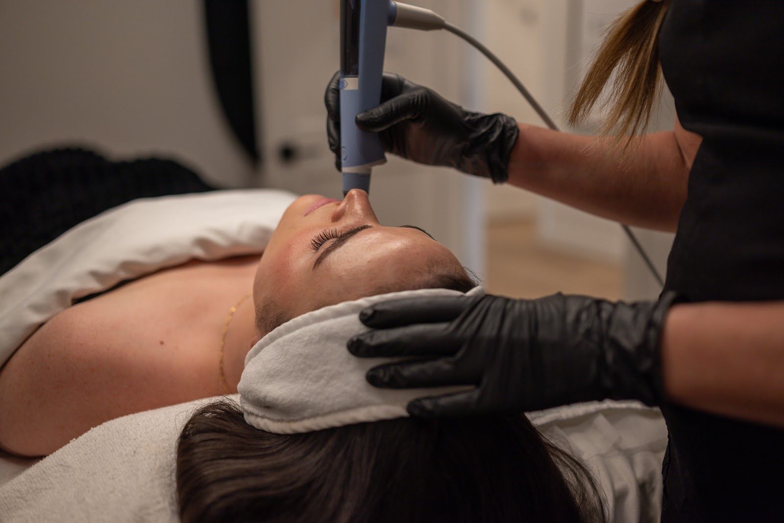 Person receiving facial treatment with tool. Esthetician wearing black gloves. Spa setting.