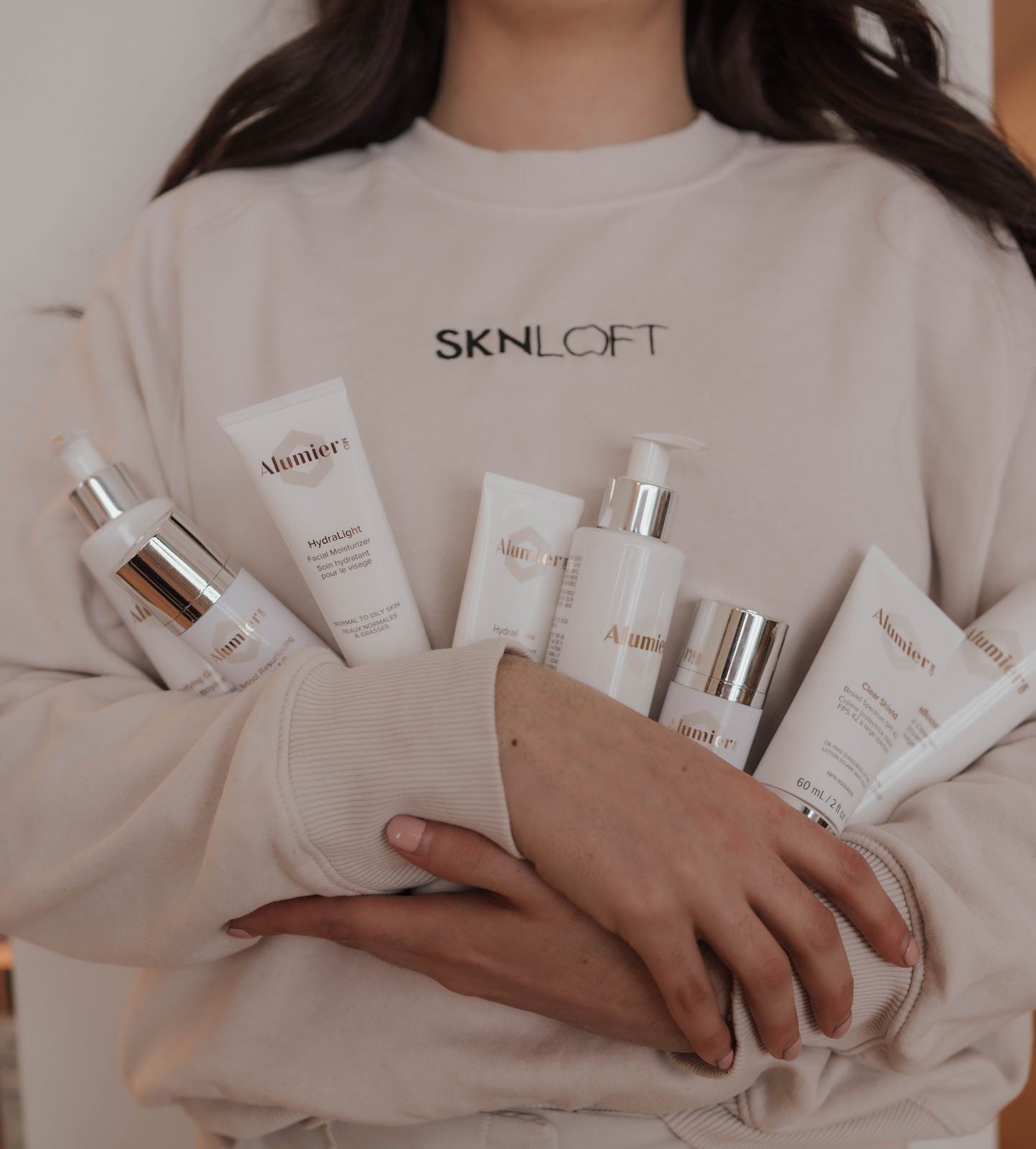 Person holding various white skincare product bottles. SKNLIFT logo on sweatshirt.