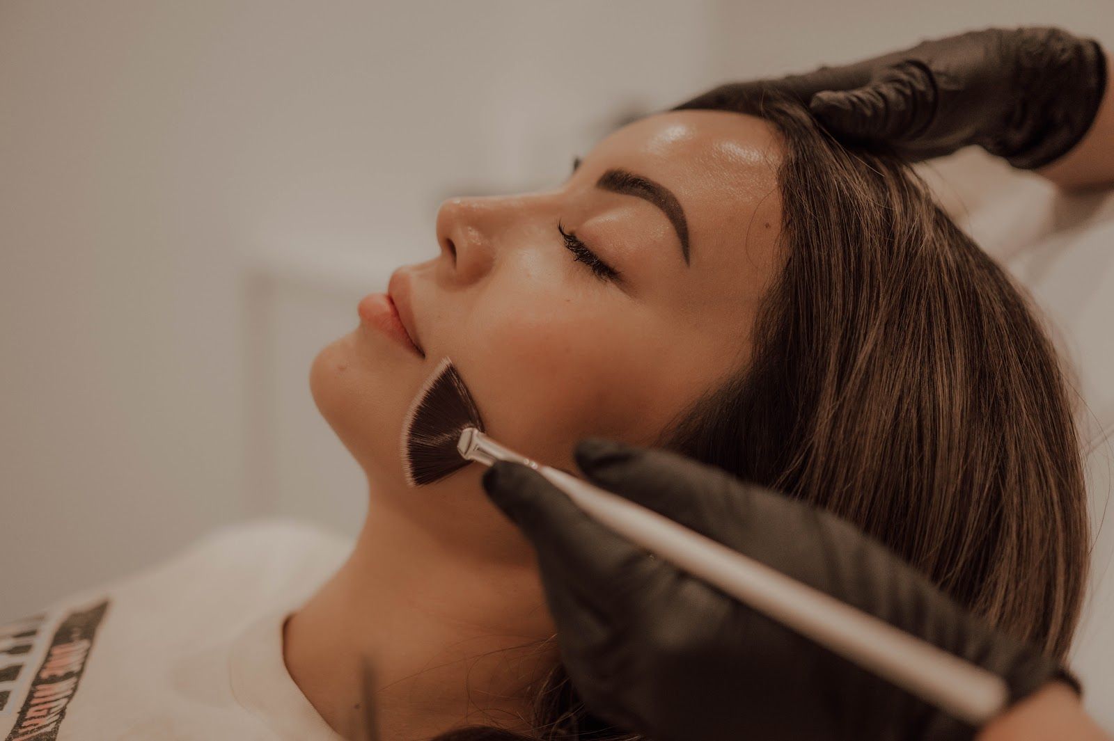 Person receiving facial treatment with a brush. The person has closed eyes, with gloved hands applying a product.