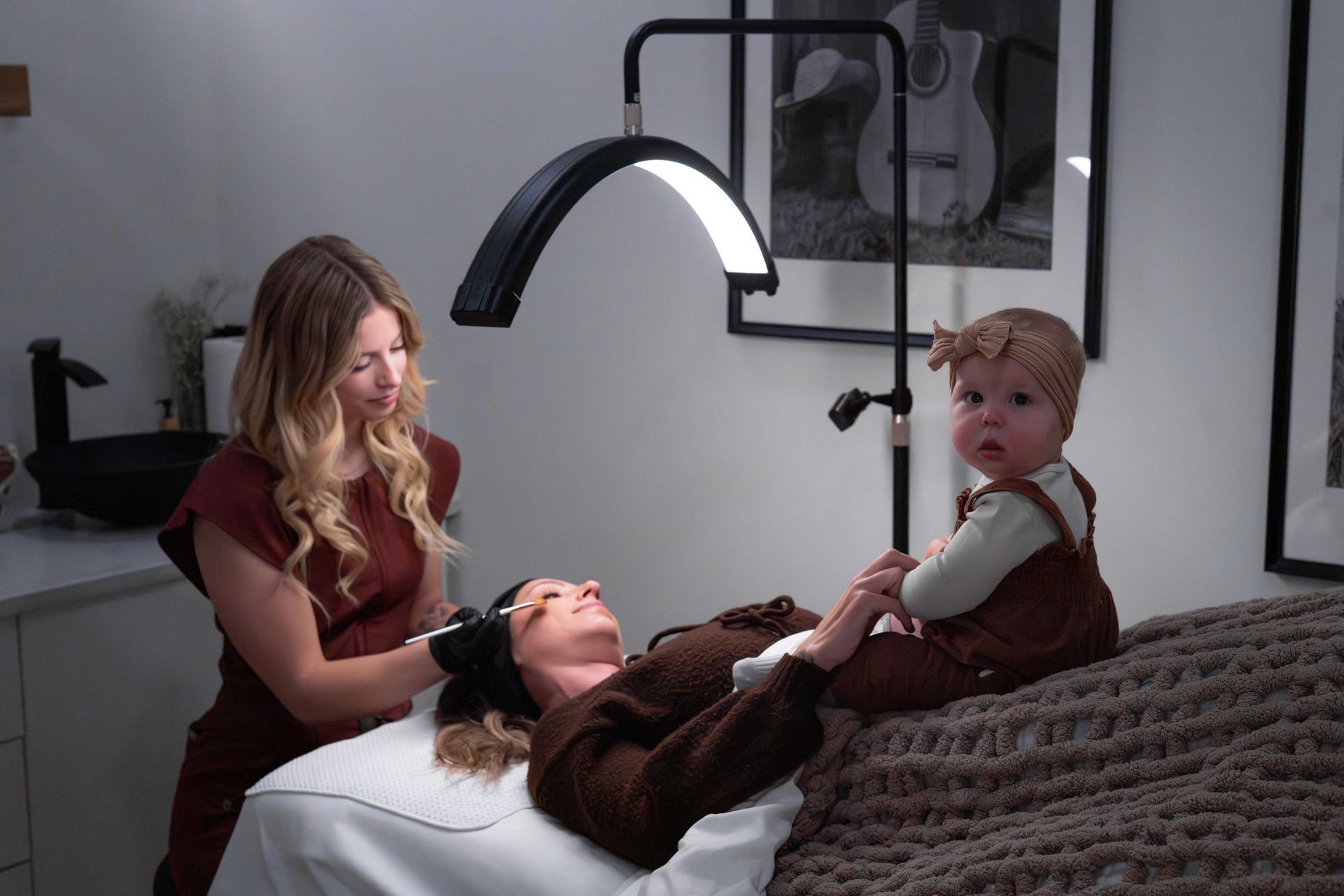 A technician performs a facial treatment on a client lying on a table, with a small child sitting nearby under a lamp.
