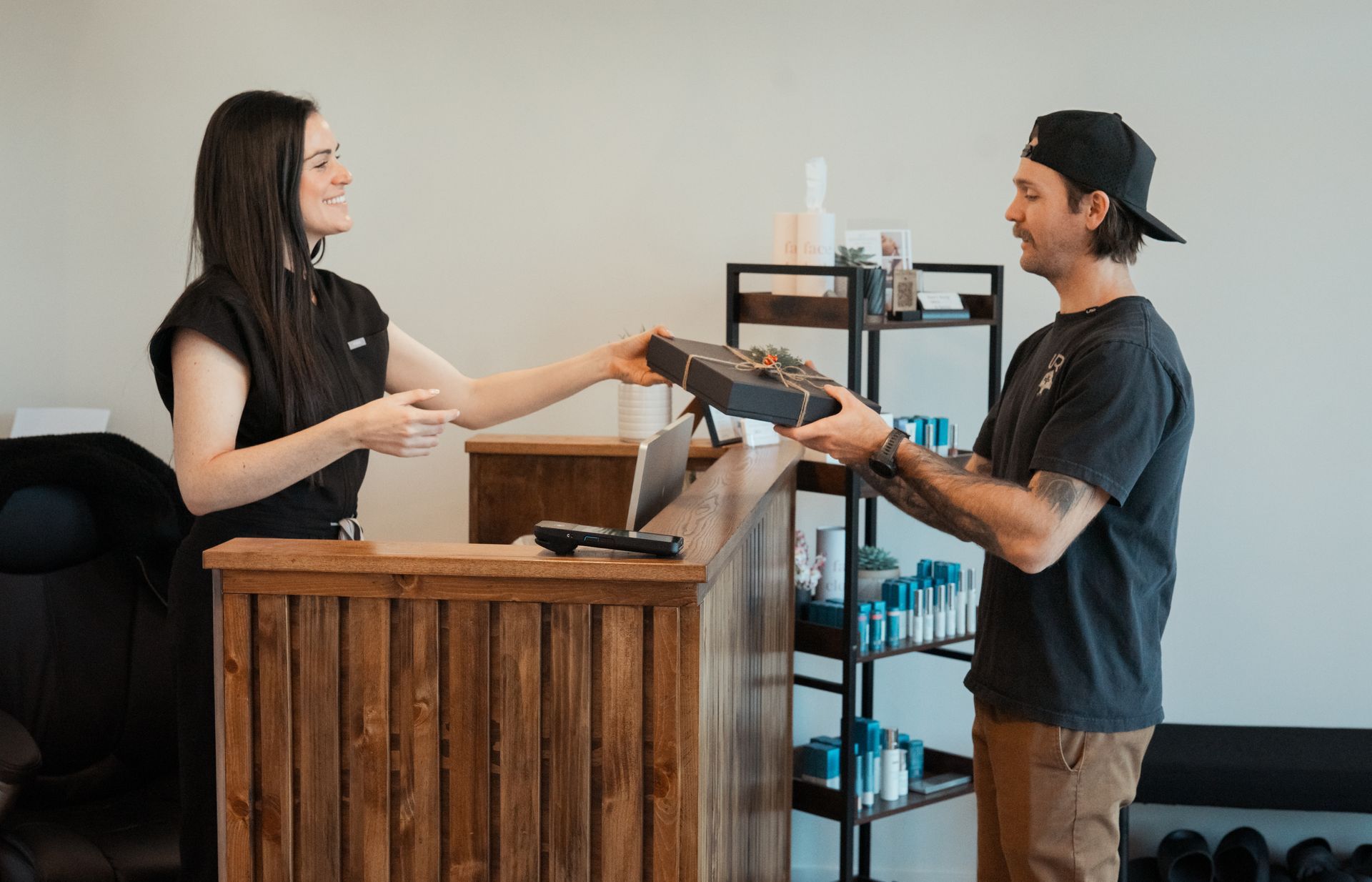 An employee hands a black gift box to a customer across a wooden reception counter in a shop.