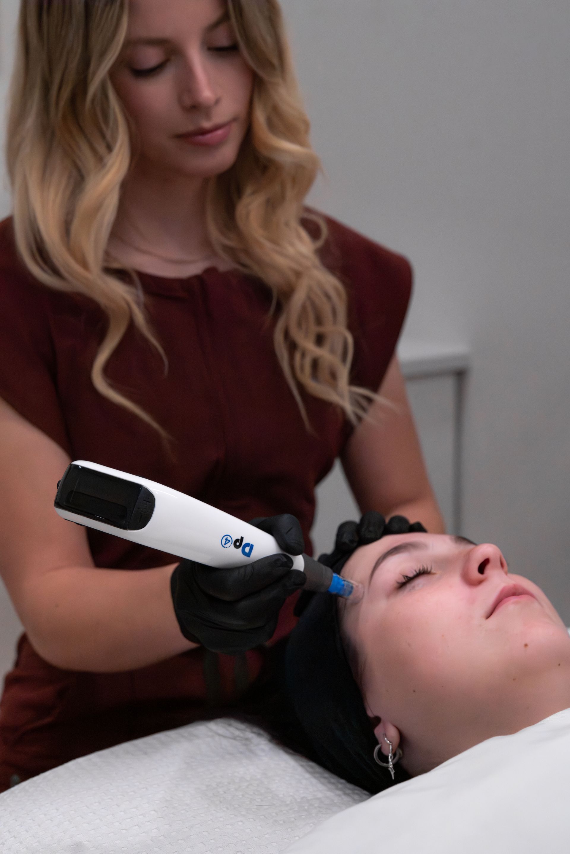 Woman receiving a cosmetic procedure on her face with a hand-held device. Another woman holds the device. Indoors, neutral setting.