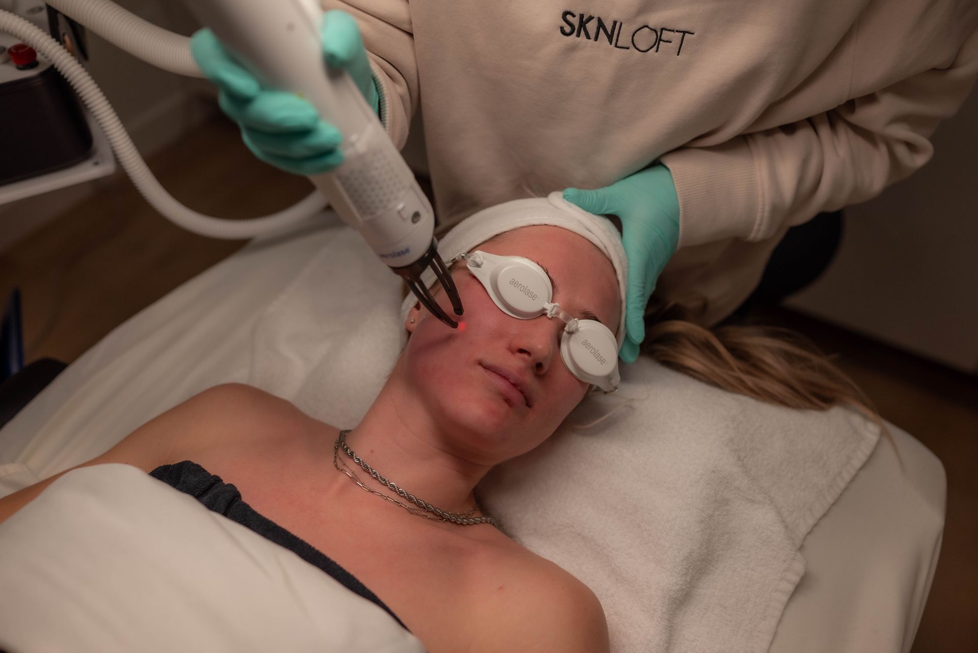 Woman receiving facial laser treatment at a salon. Operator holds device over red-marked skin.