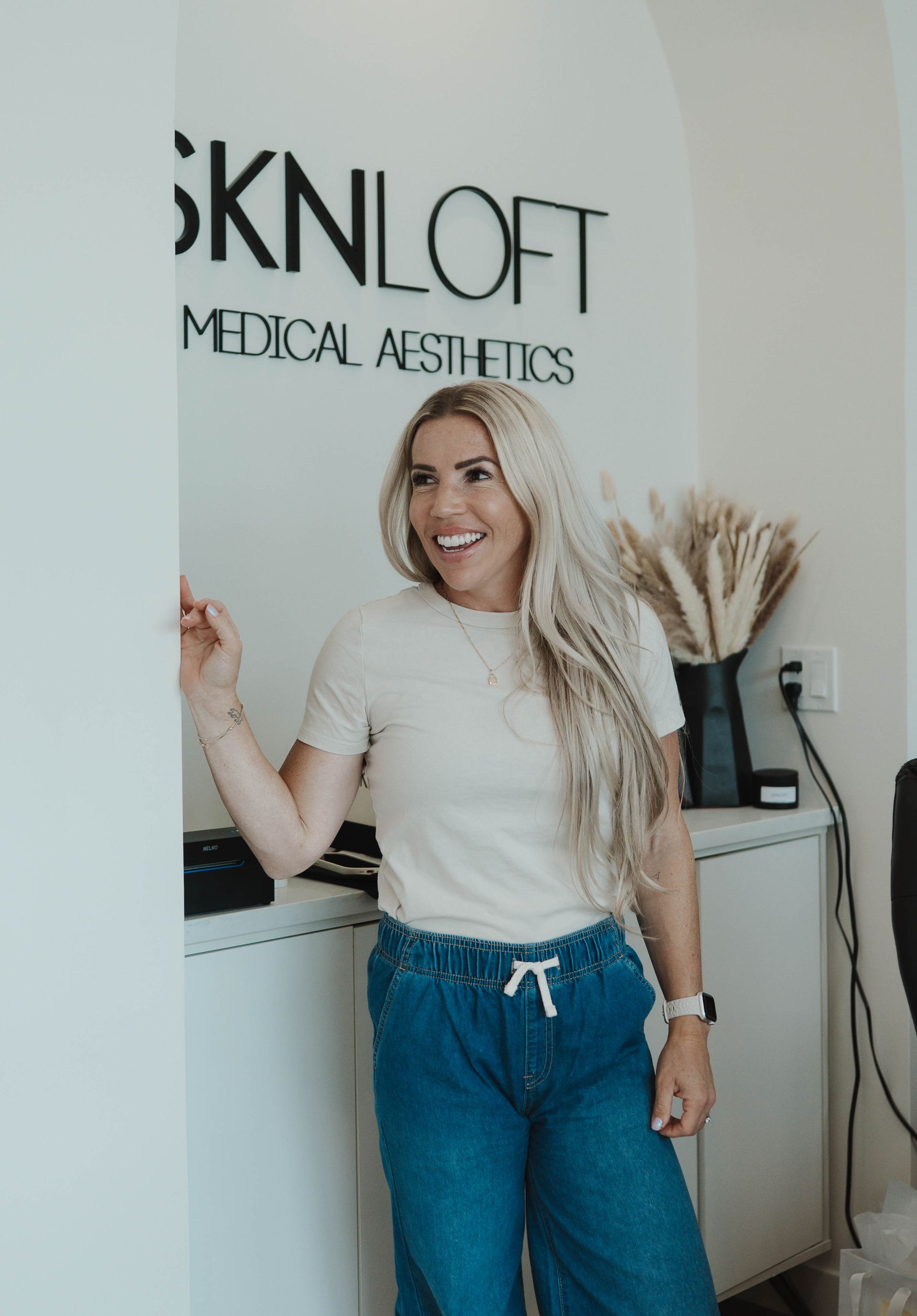 Woman smiling, leaning on a white wall in a medical aesthetics office,