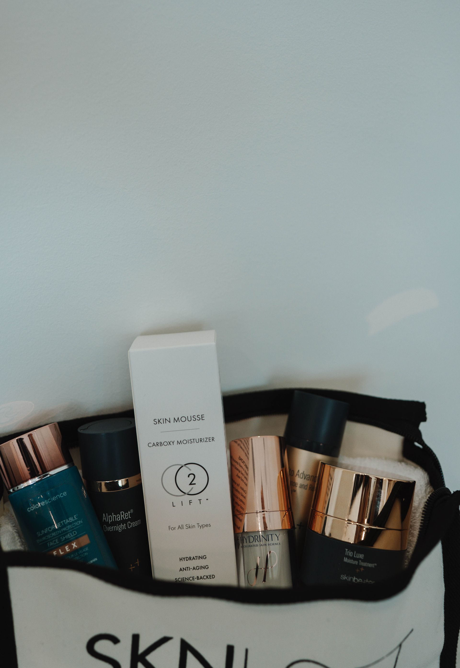 Cosmetic products in a SKNLY branded bag against a white wall.