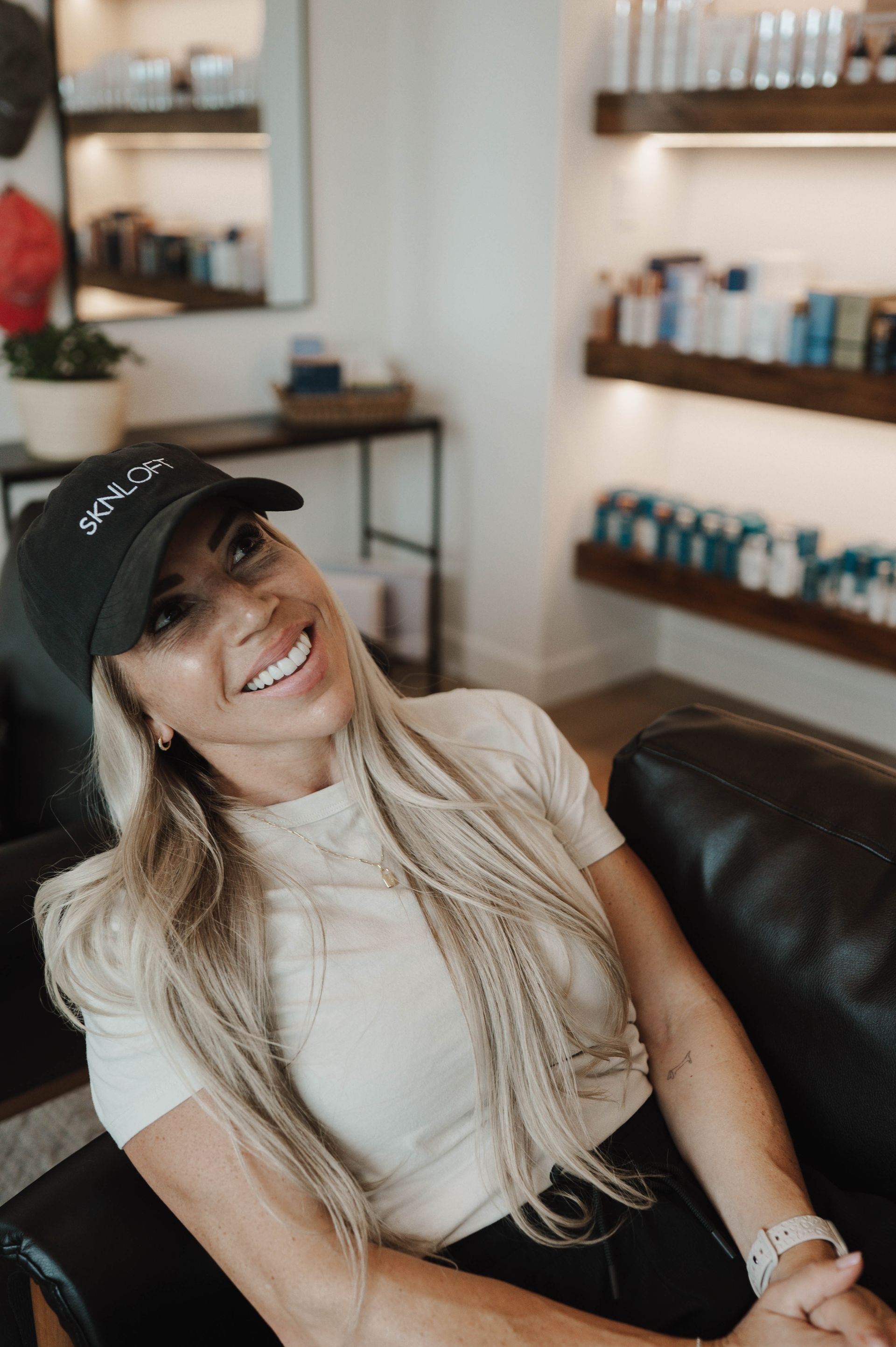 Woman wearing a hat smiles while seated in a salon, looking upward. Shelves of products are in the background.