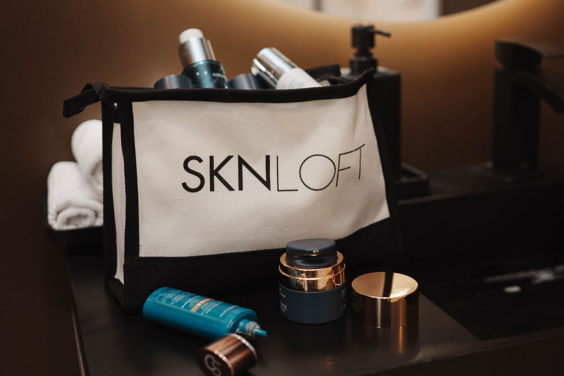 Cosmetic products and SKN LOFT bag on a bathroom counter with towels and a faucet.