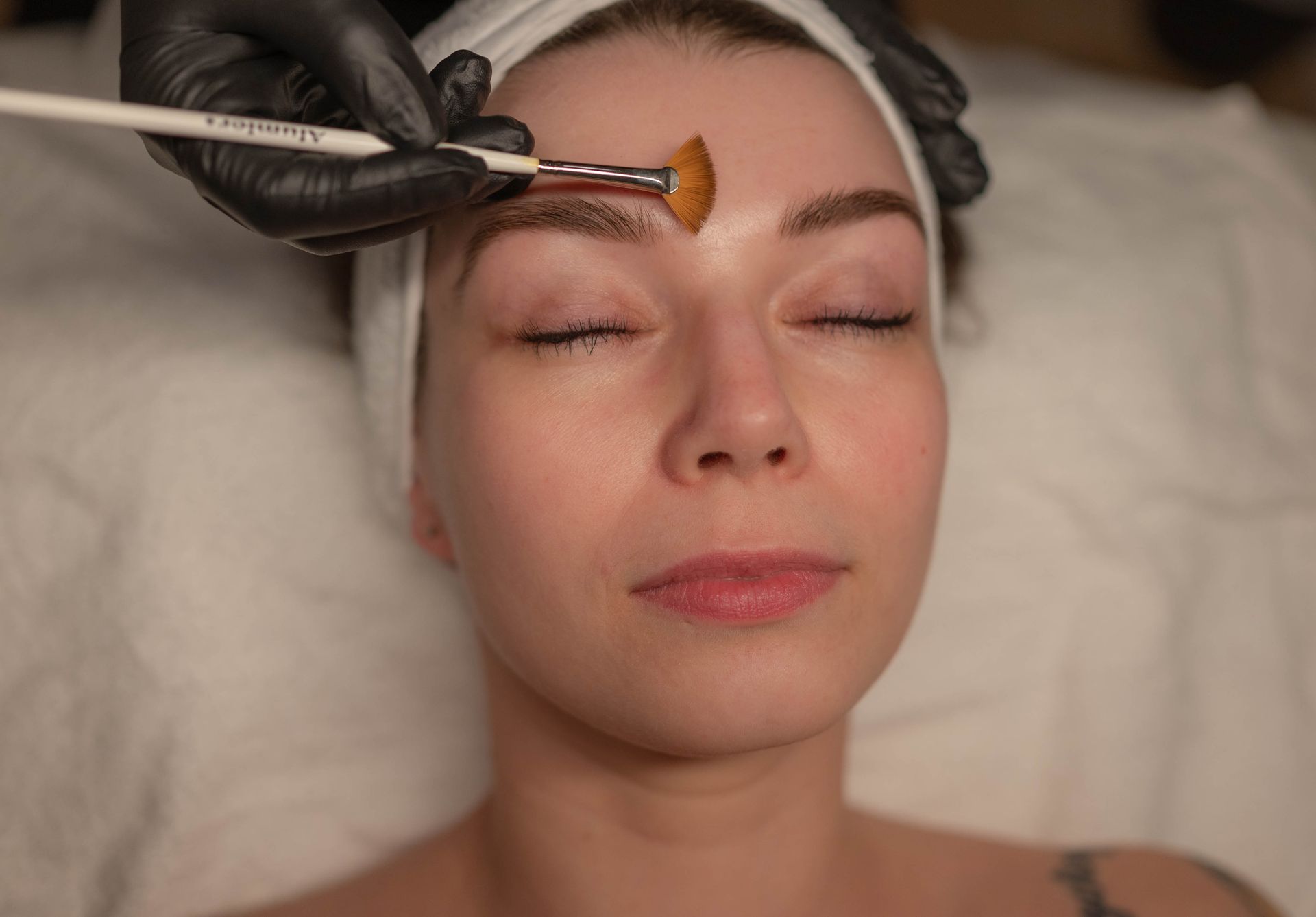Woman receiving a facial peel, eyes closed, with a brush applying a product on her forehead.