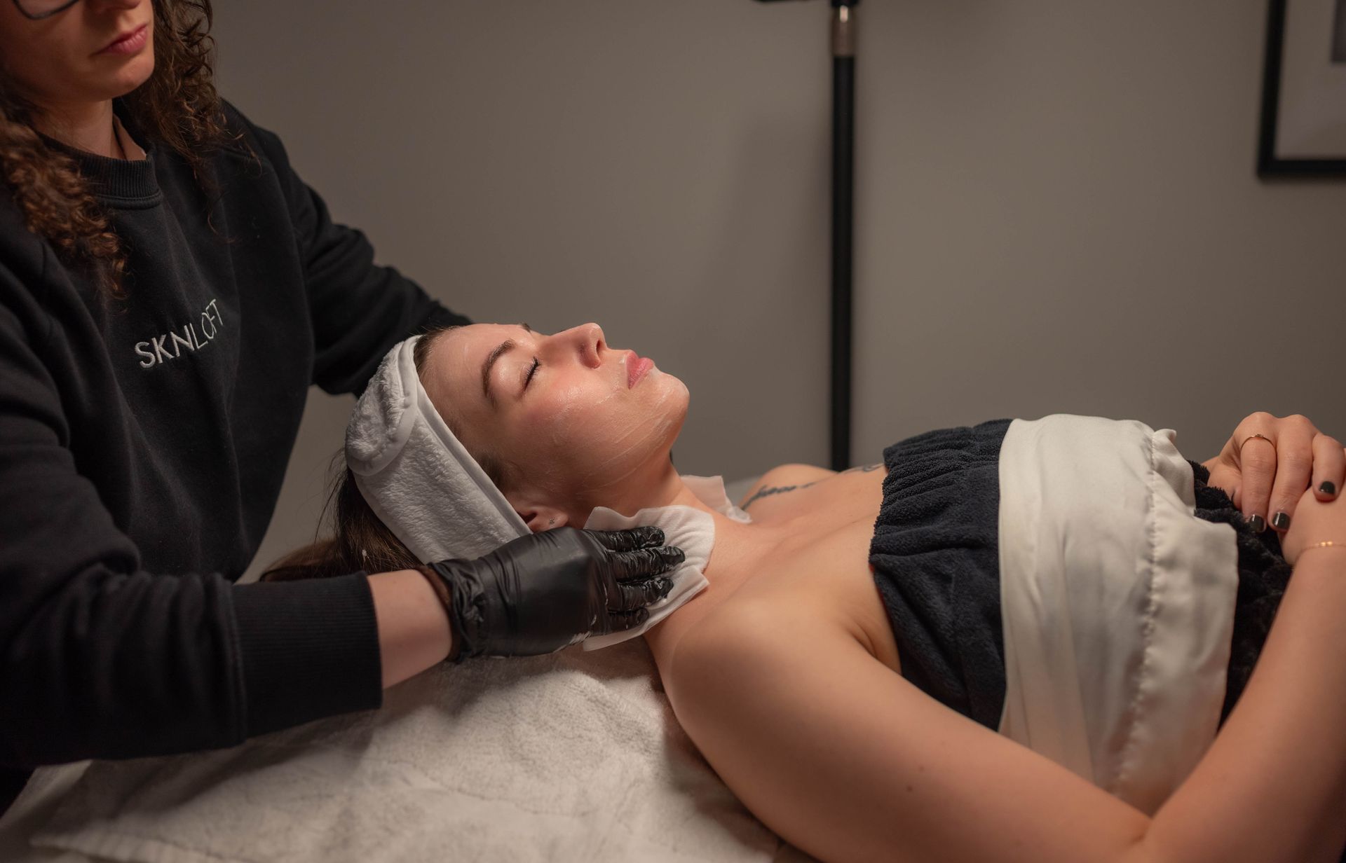 Woman receiving facial treatment at a spa; therapist in black gloves wiping her face, white towel on head.