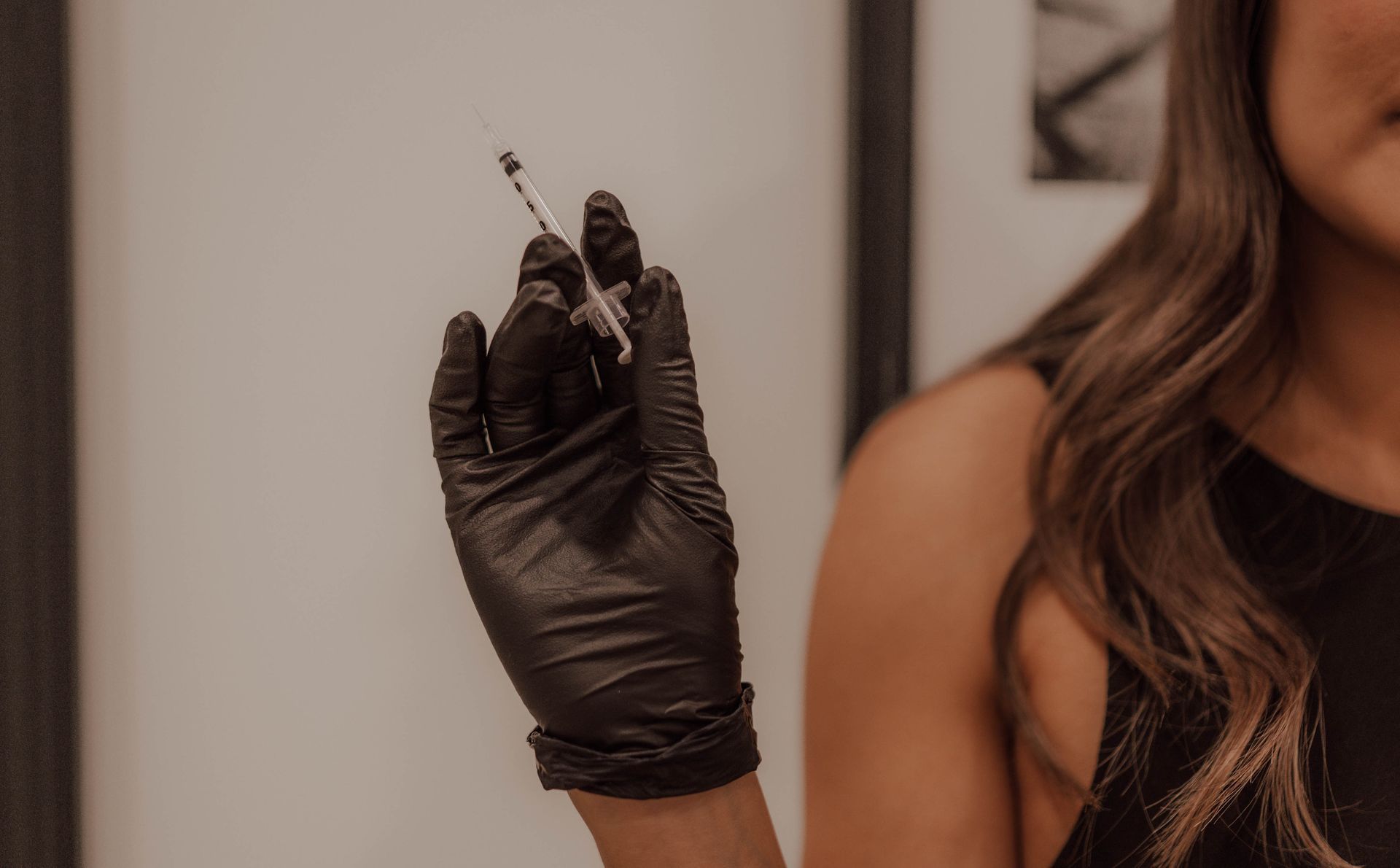 A hand in a black latex glove holding a small syringe, with a person visible in the background.