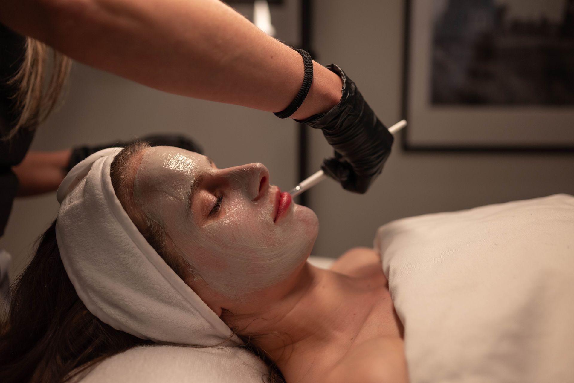 A person receiving a facial treatment with a clay mask applied by an esthetician using a brush.