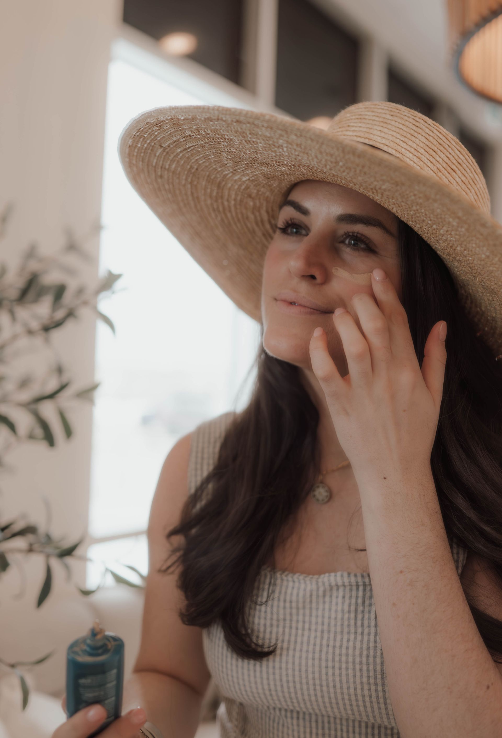 A woman wearing a straw hat is applying sunscreen to her face.