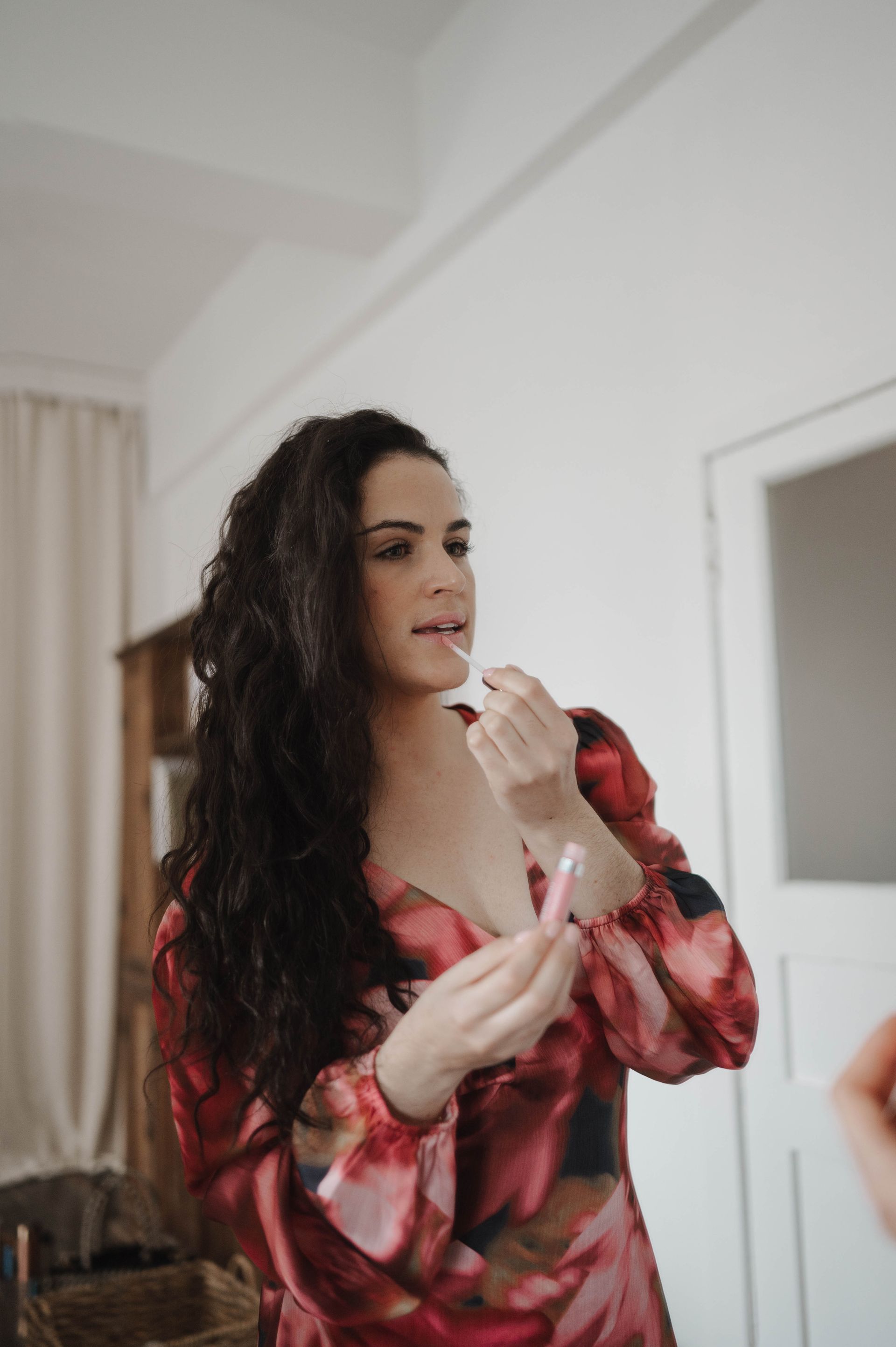 A woman in a red dress is applying lipstick in front of a mirror.