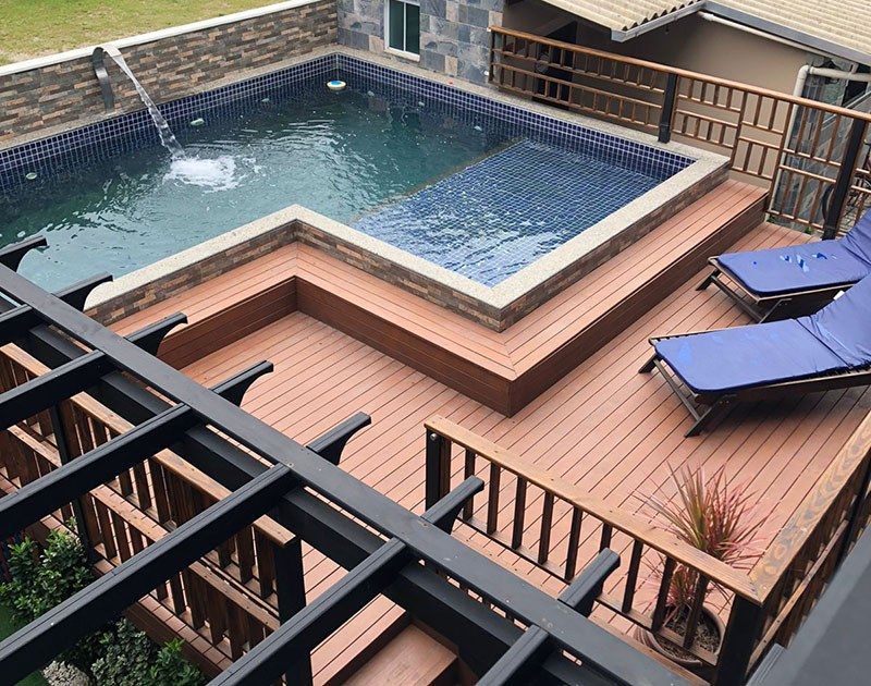 Um terraço elevado ao ar livre com piscina revestida de azulejos, uma fonte e duas espreguiçadeiras azuis sob uma pérgola.