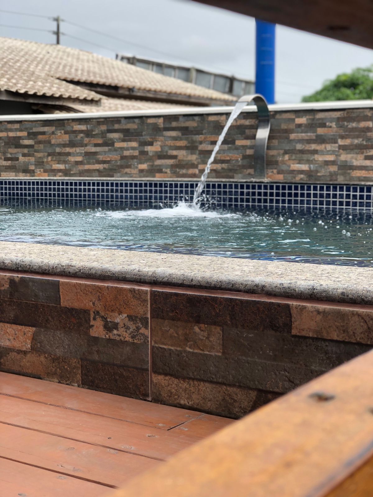 Uma fonte de aço inoxidável deságua em uma piscina no quintal, revestida com azulejos de mosaico azul e paredes de pedra marrom.