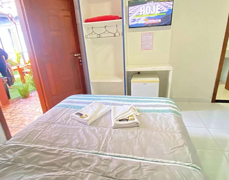 Um quarto de hotel arrumado com uma cama bem feita, um pequeno guarda-roupa aberto, uma televisão e um frigobar ao lado de uma porta de madeira.