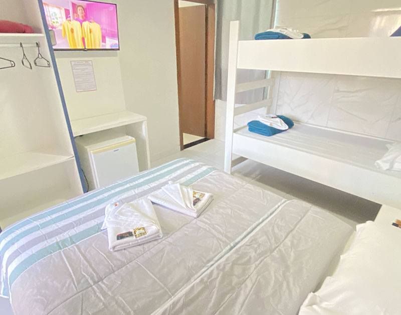 Um quarto de hotel com uma cama de casal, um beliche branco, um frigobar e uma televisão na parede.