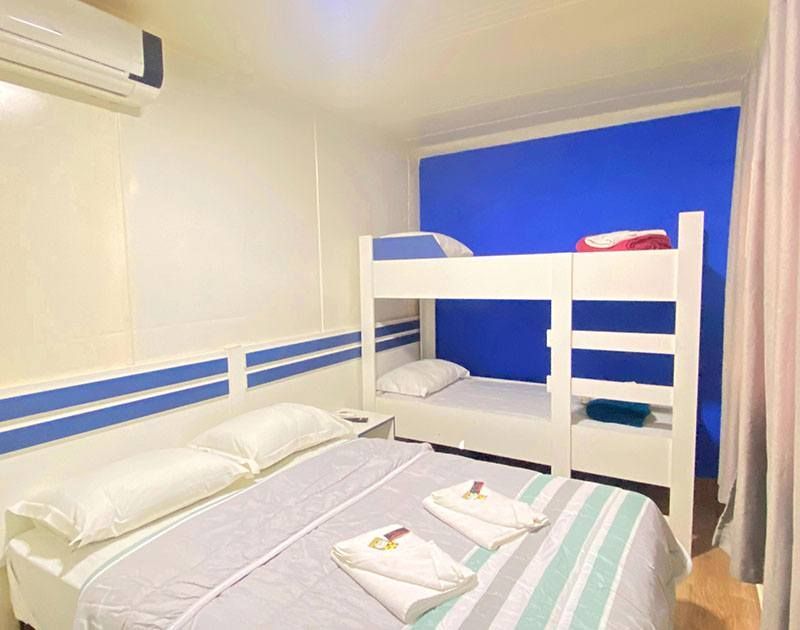 O quarto possui uma cama de casal com lençóis brancos e um beliche branco encostado em uma parede de destaque azul vibrante.