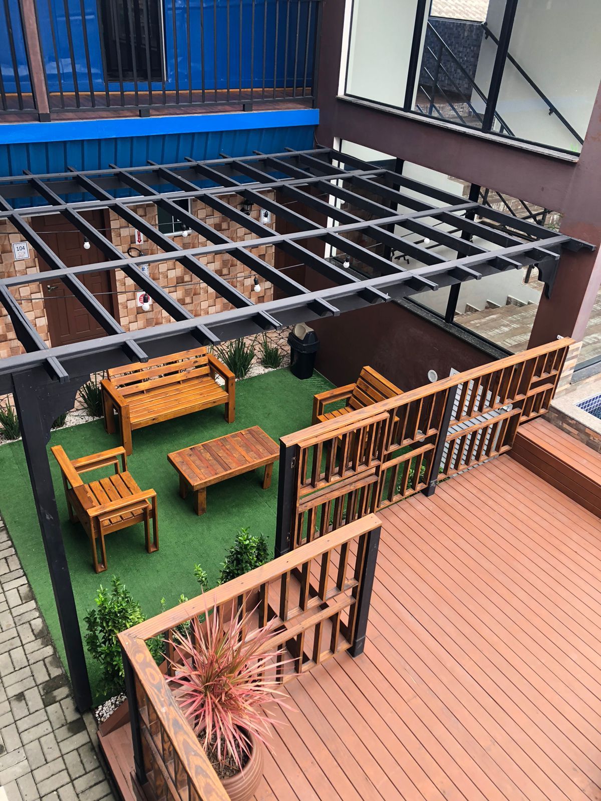 Uma área de pátio nos fundos com uma pérgola de madeira sobre um gramado com móveis de madeira, ao lado de um deck com corrimão.