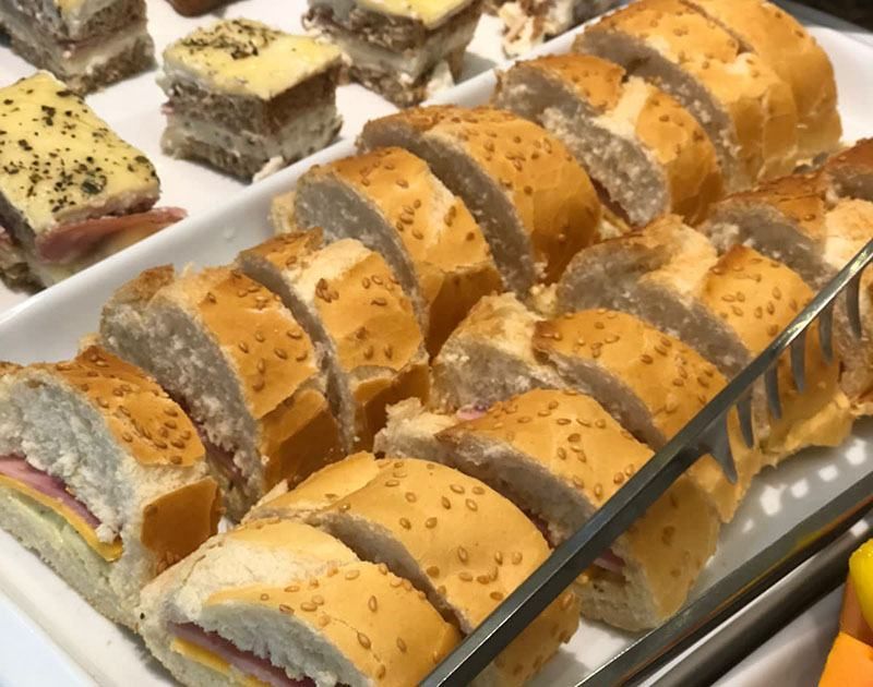 Um close de uma bandeja repleta de diversos sanduíches, incluindo sanduíches de chá cortados em quadrados e pãezinhos de gergelim fatiados.