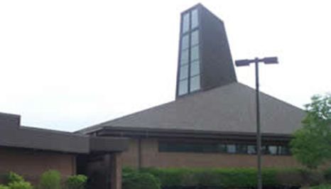 Church building with a dark steeple featuring a cross-shaped window and brown roof.