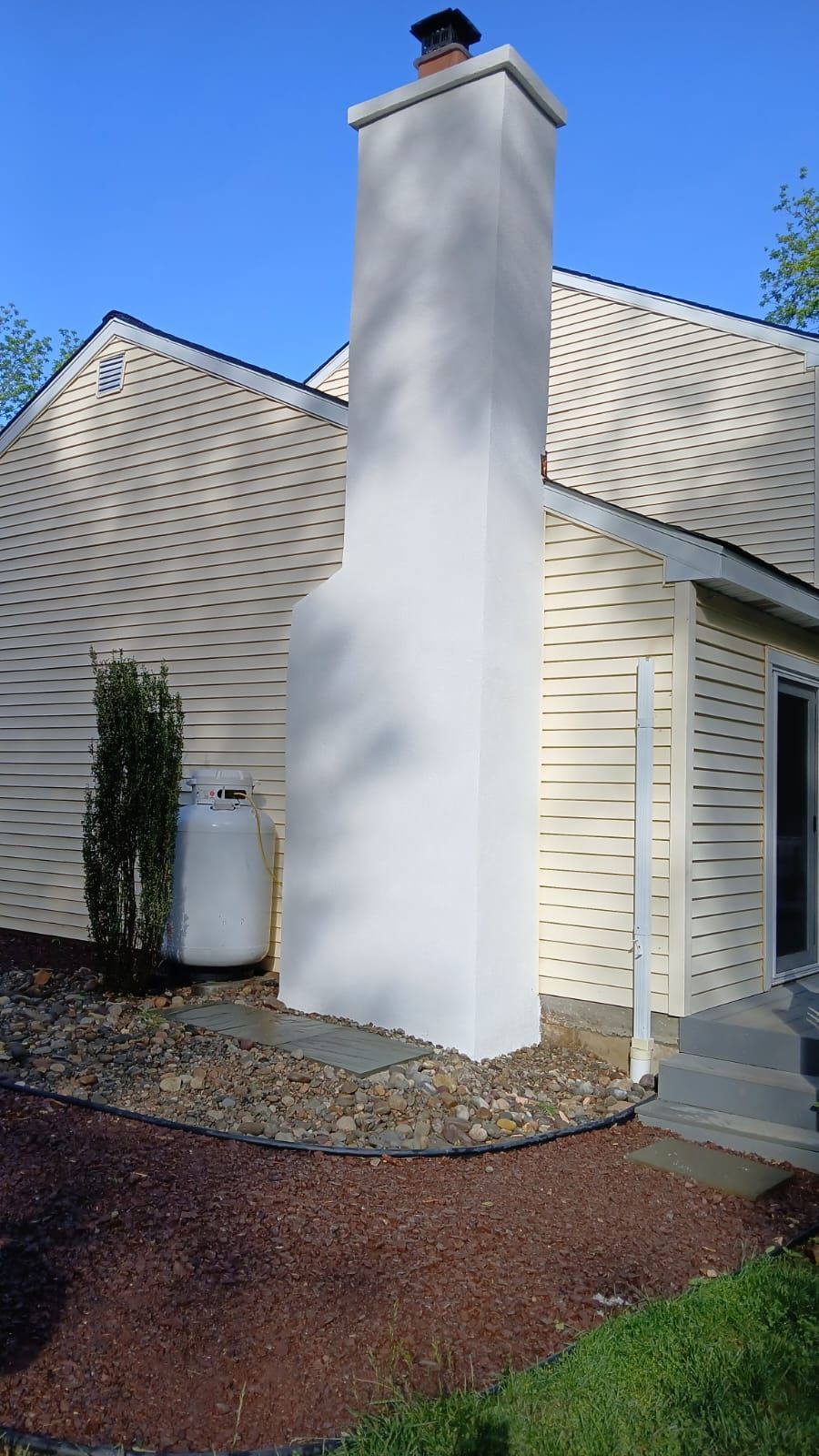 A white stucco chimney stands against the side of a house with beige siding and a gravel landscape.