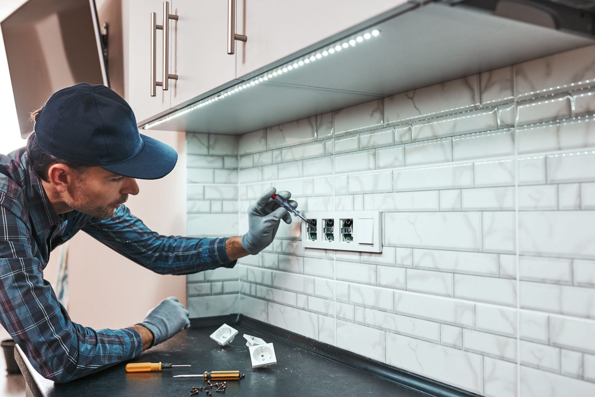 Man in cap and gloves installing electrical outlets on a tiled kitchen wall.