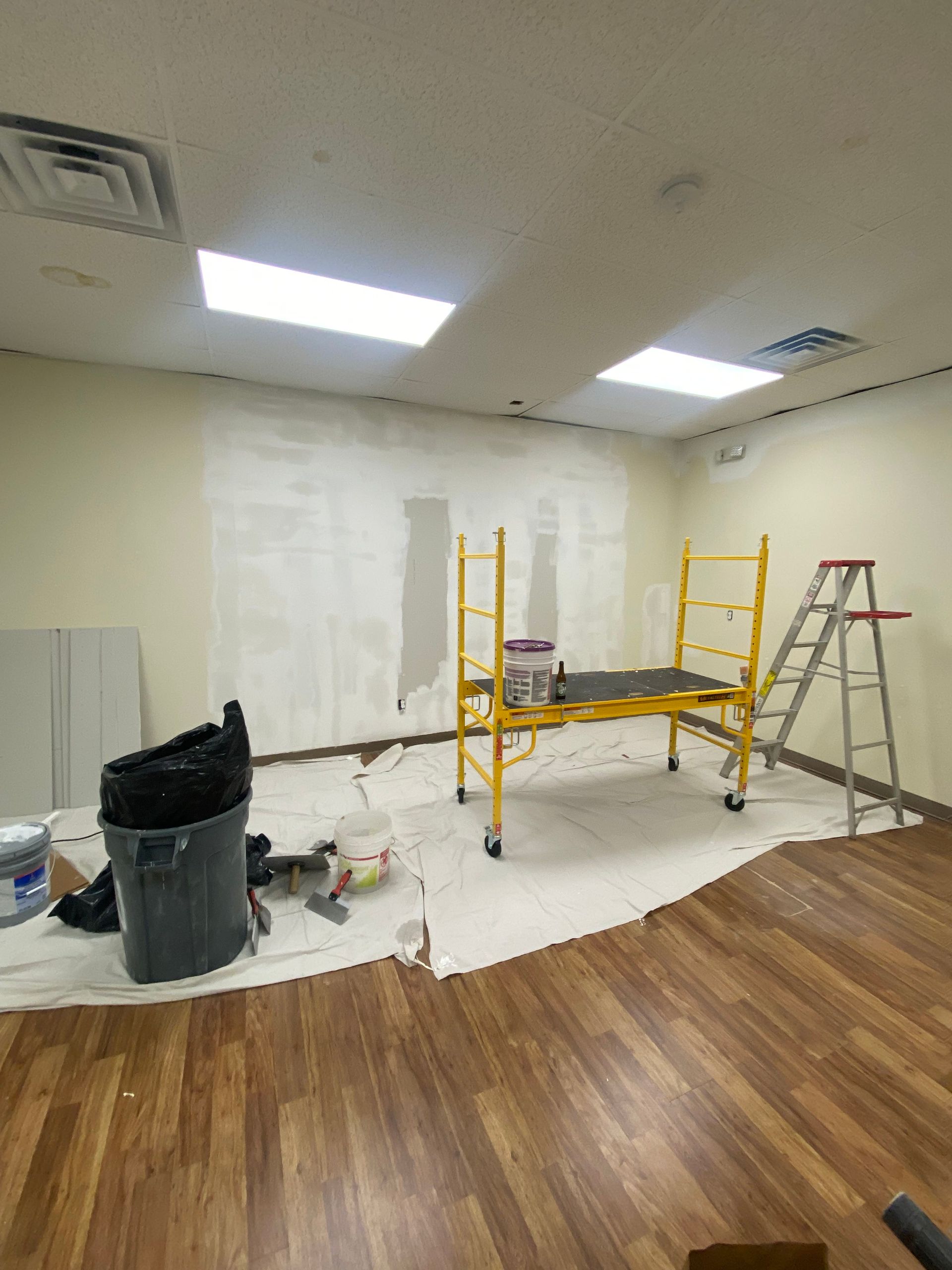 A room under renovation with paint on the wall, scaffolding, drop cloths, and a trash bag.