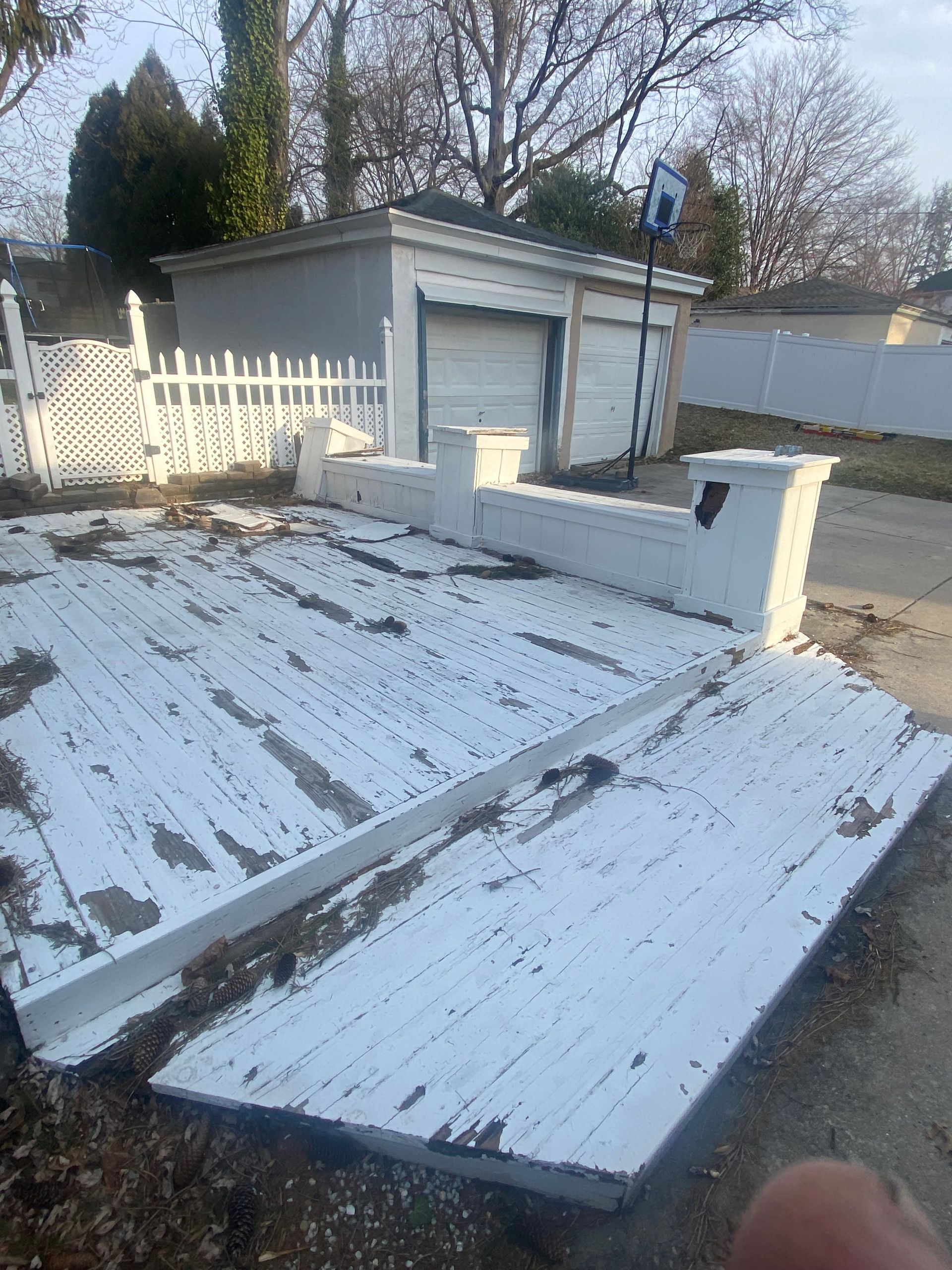 White, damaged deck in front of a white garage with a basketball hoop. A white picket fence borders the left.