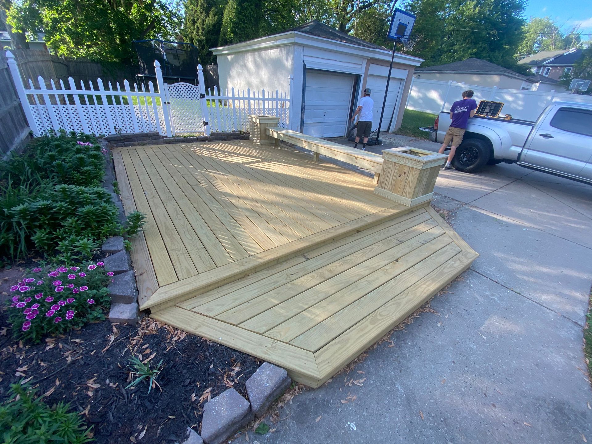 Newly built wooden deck in a front yard, two workers nearby, a white garage in the background, and a white fence to the left.