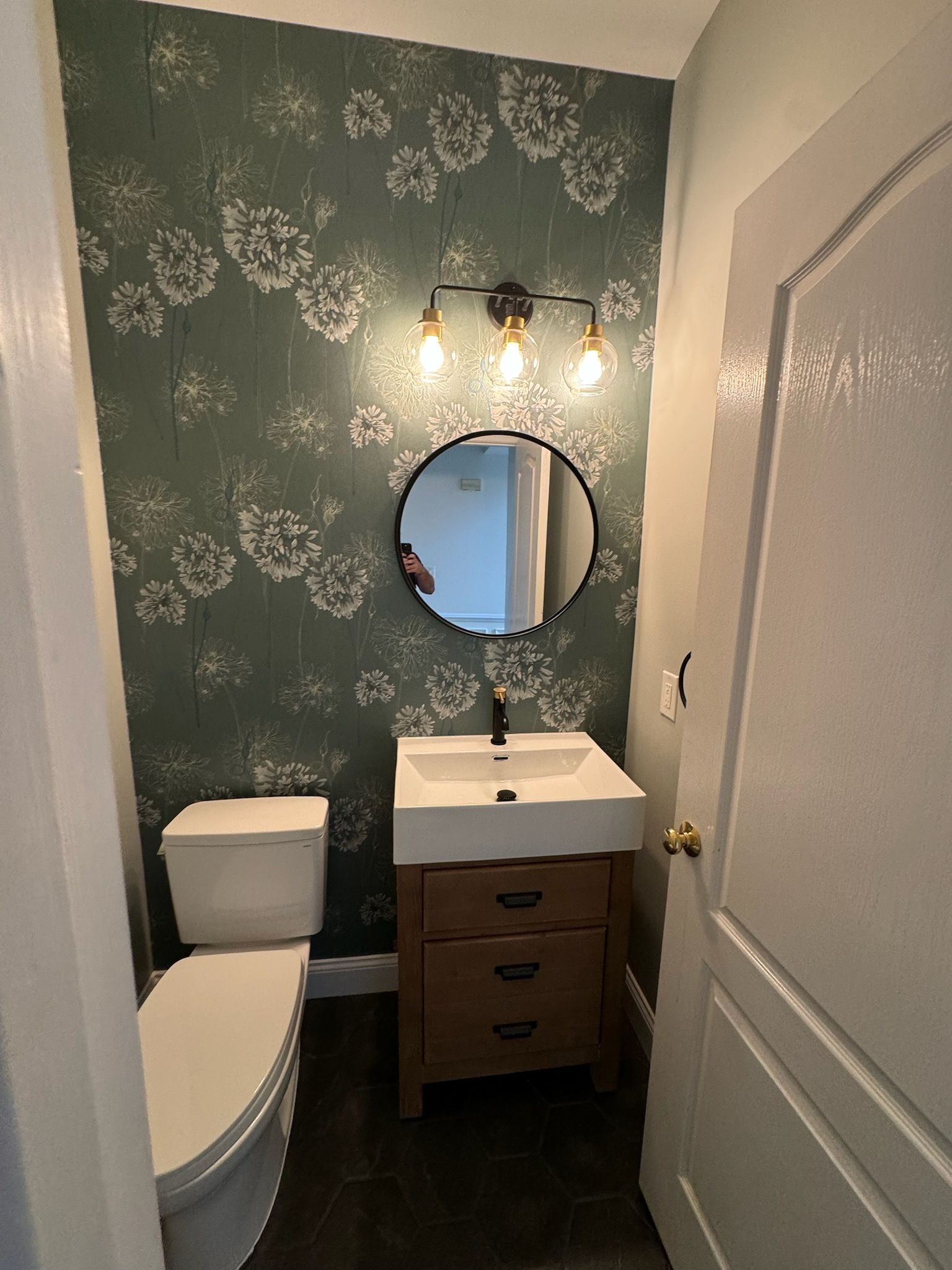 Small bathroom with floral wallpaper, wood vanity, round mirror, and a toilet.