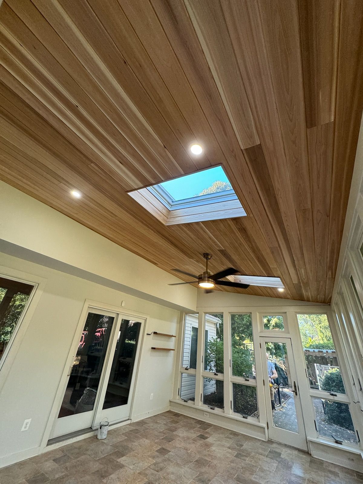Sunroom with wood ceiling, skylight, and ceiling fan. Glass doors and windows overlook a green yard.