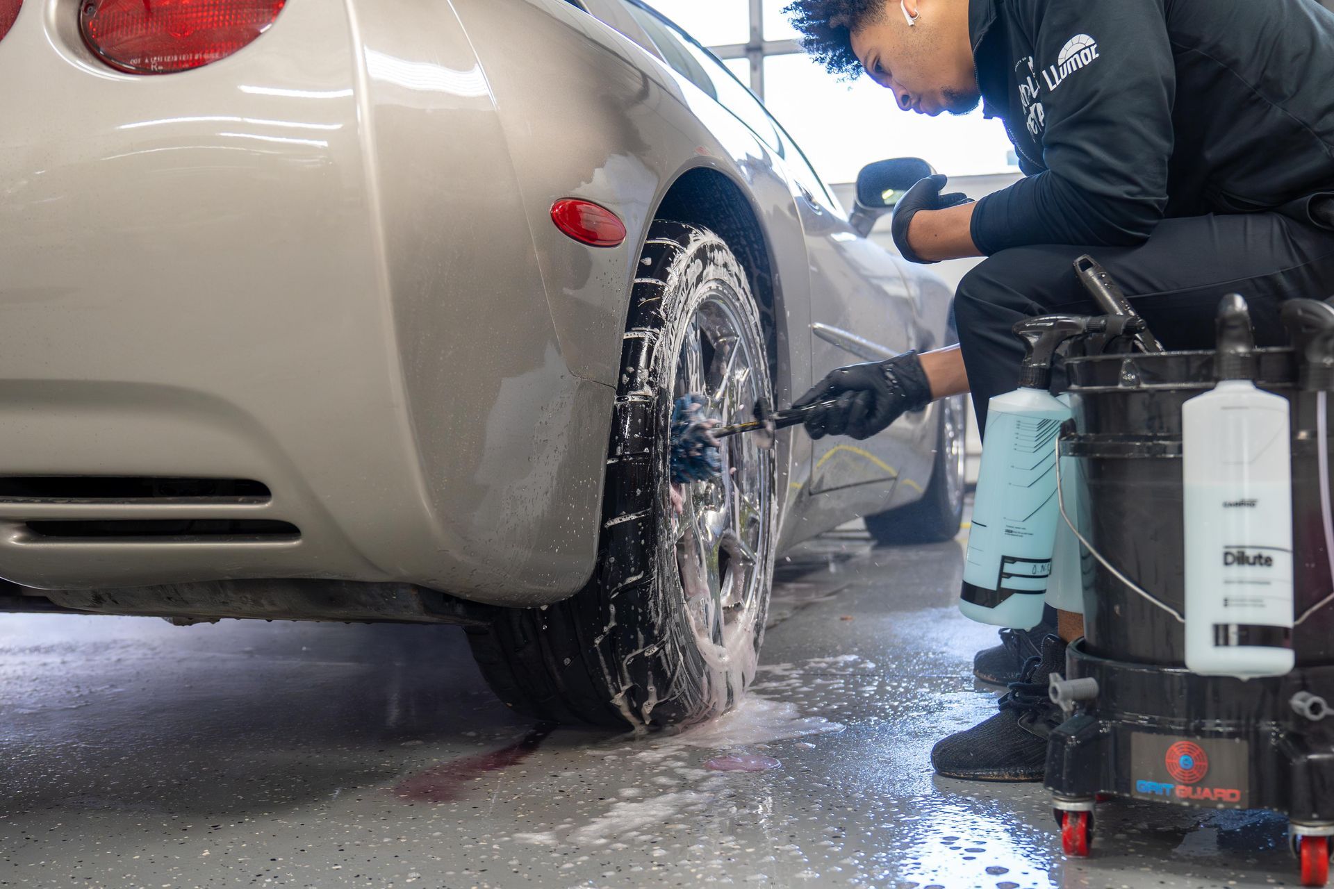 Person washes a silver car's tire with soap, using spray bottles and a bucket.