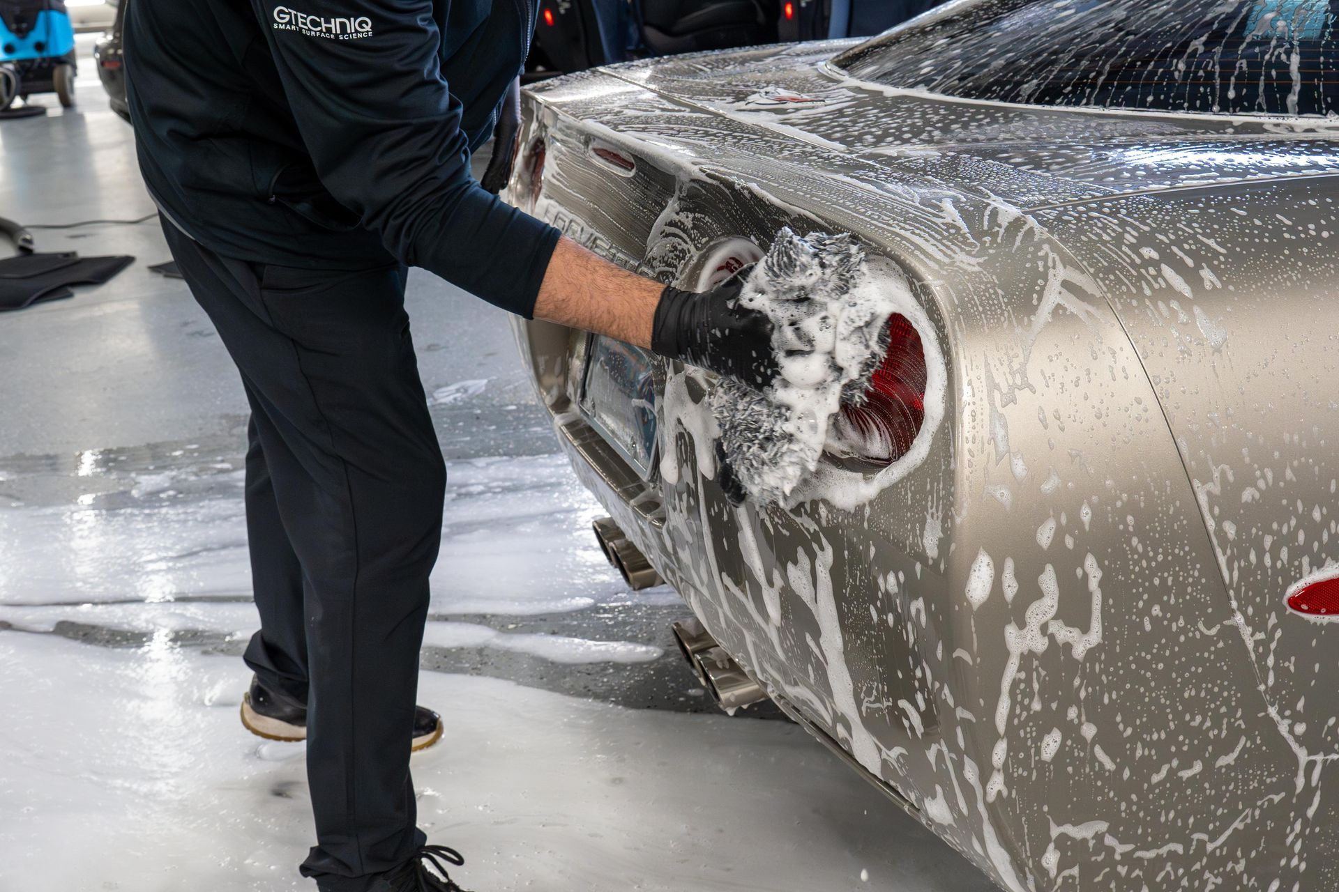 A worker wearing gloves cleans the soapy taillight of a silver sports car in a garage.