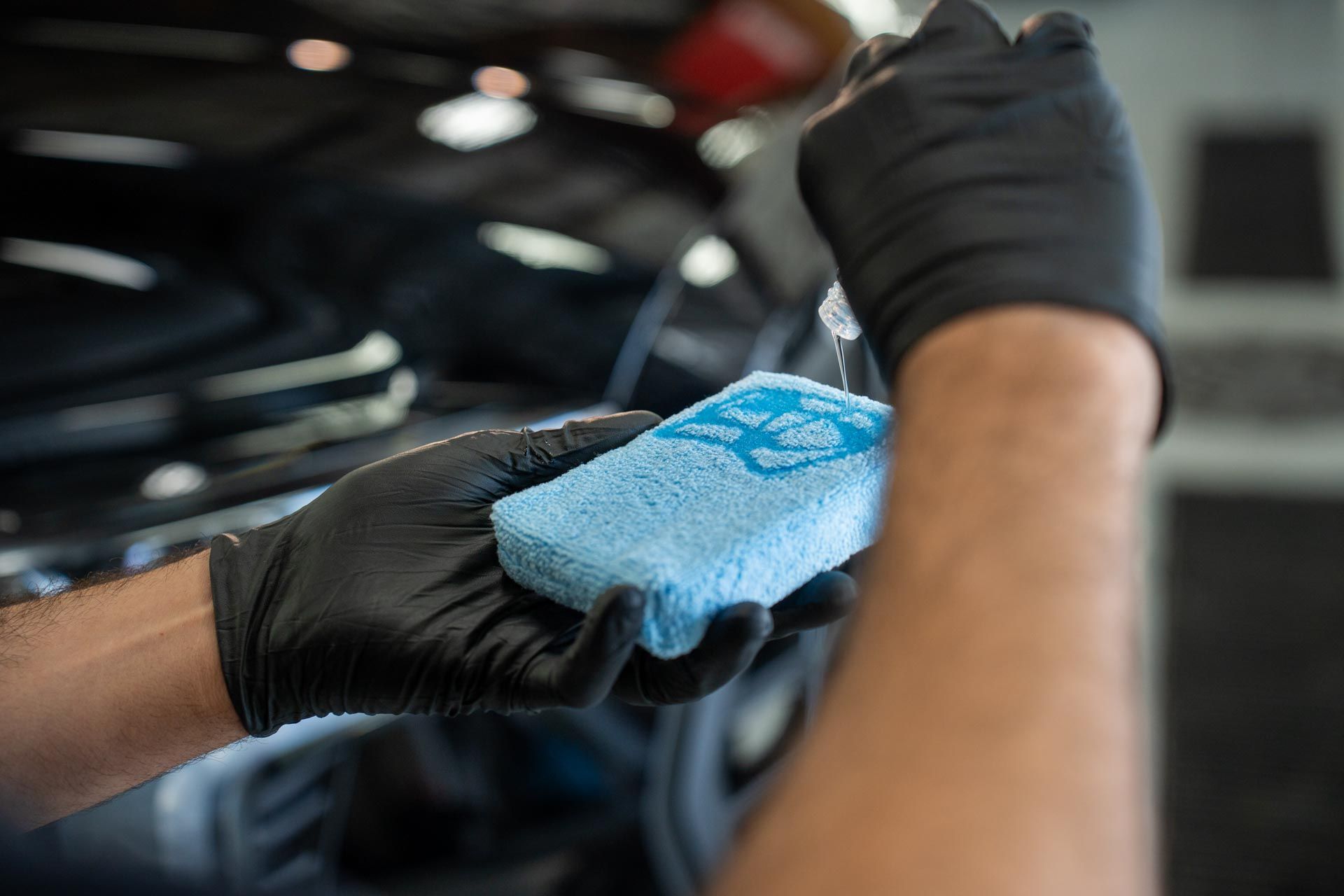 Gloved hand applying ceramic coating to a shiny blue car's hood in a garage.