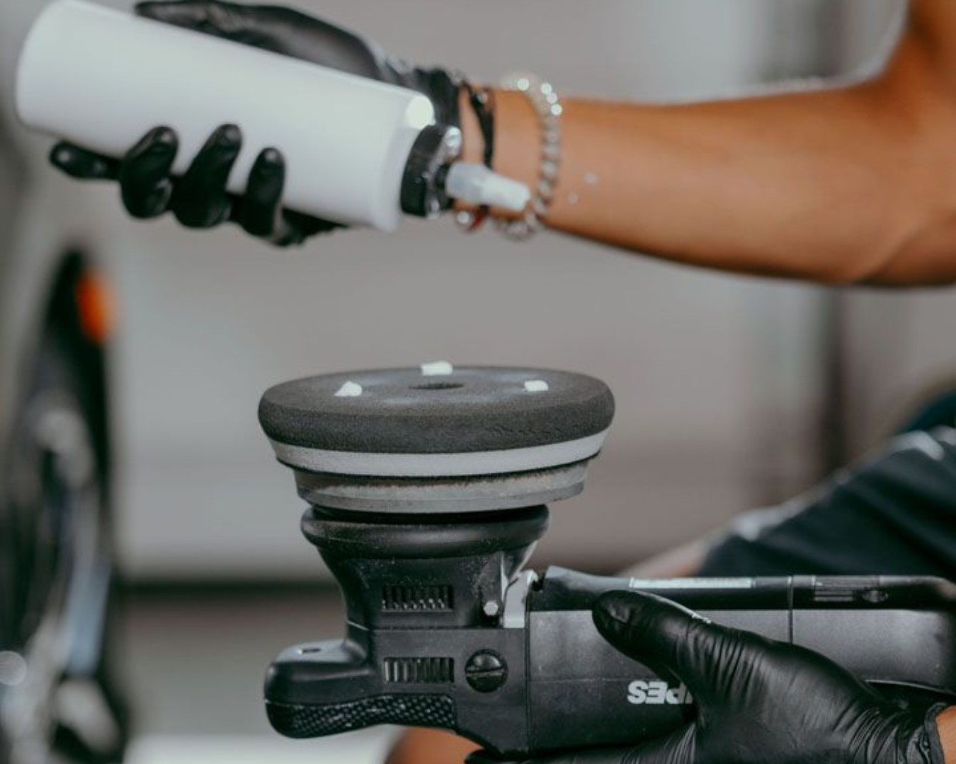 A person using a power polisher on a white car, wearing black gloves.