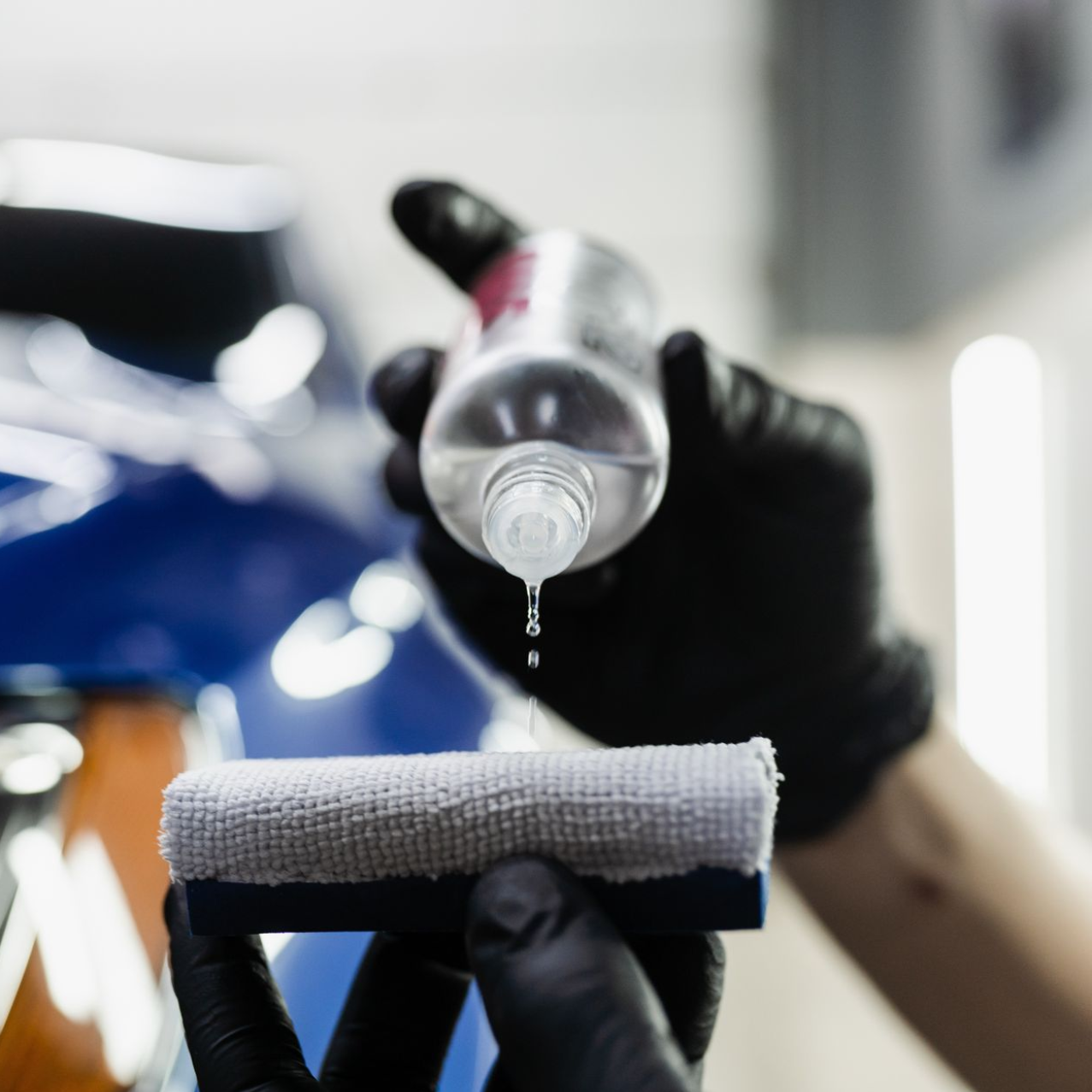 Gloved hands applying liquid from a bottle to a microfiber applicator for car detailing.