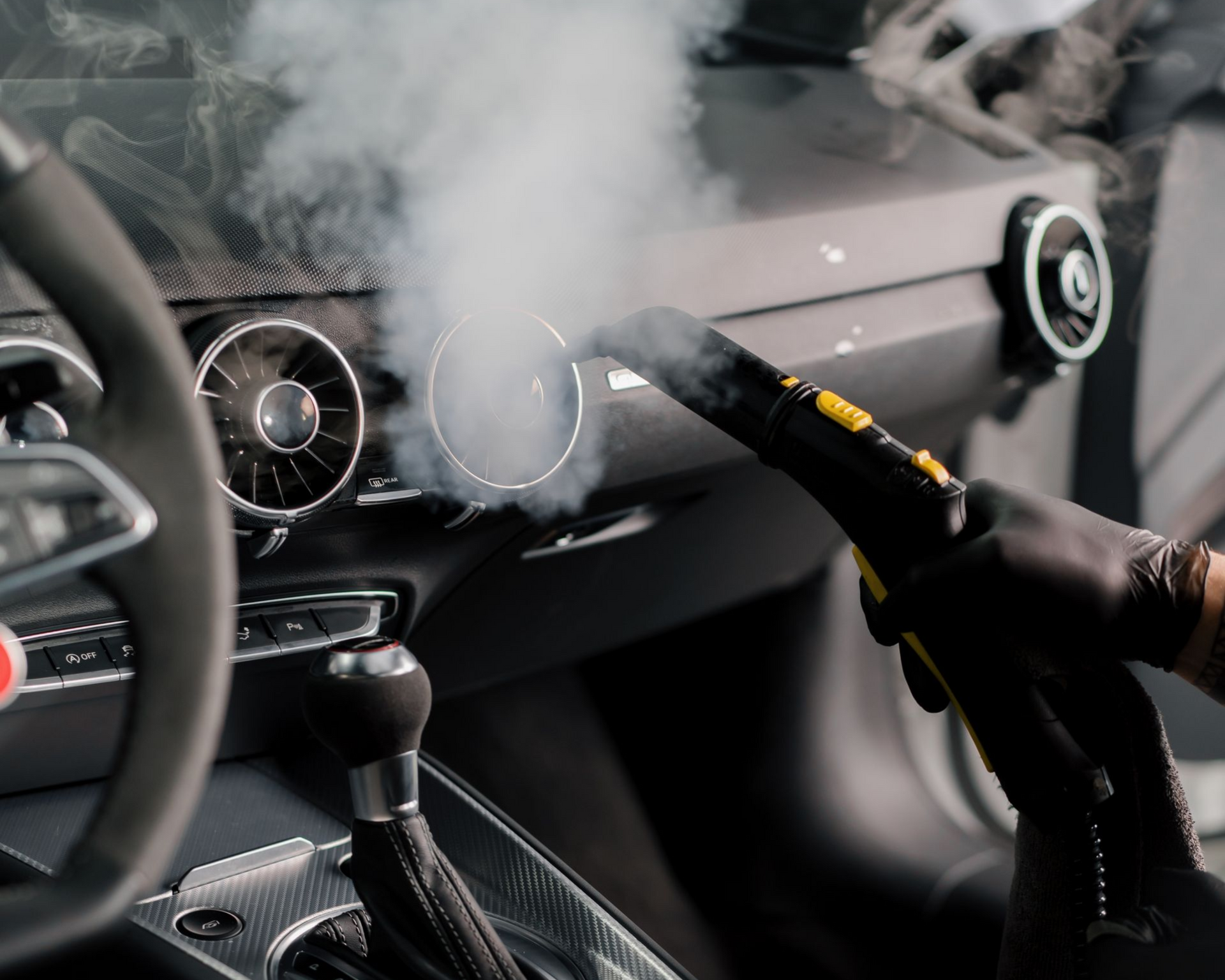 Gloved hand steaming car's dashboard vents; steam cloud visible.