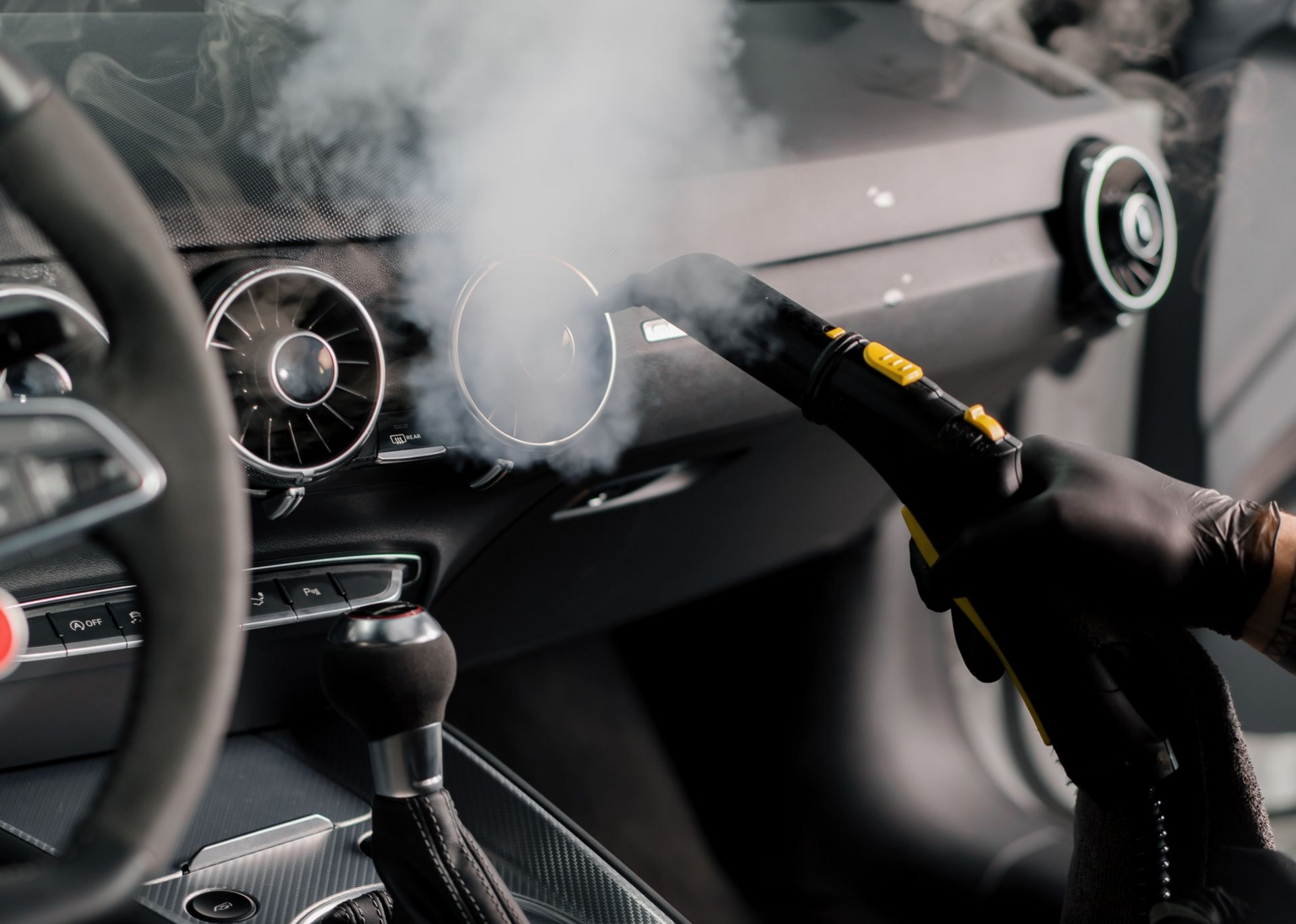 Steam cleaning car dashboard vents with a handheld device, dark interior.