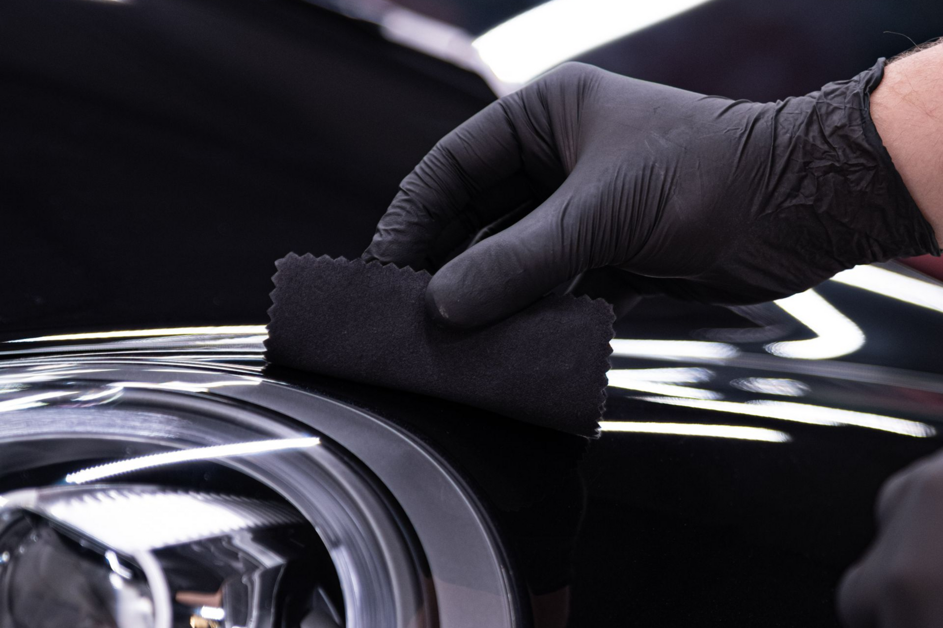 Black-gloved hand applying coating to a black car headlight, using a dark applicator sponge.