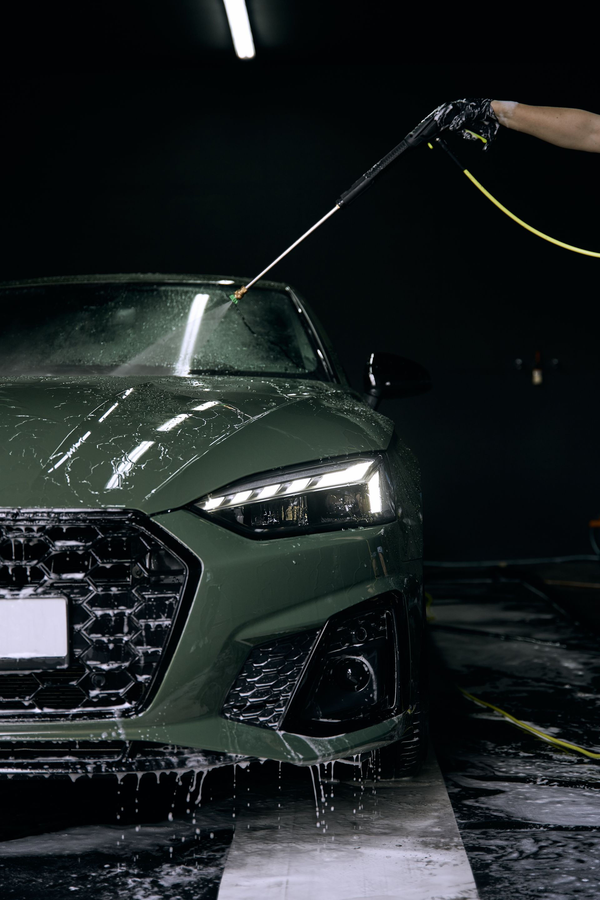 Green car being washed in a dark garage; water spraying from a hose.