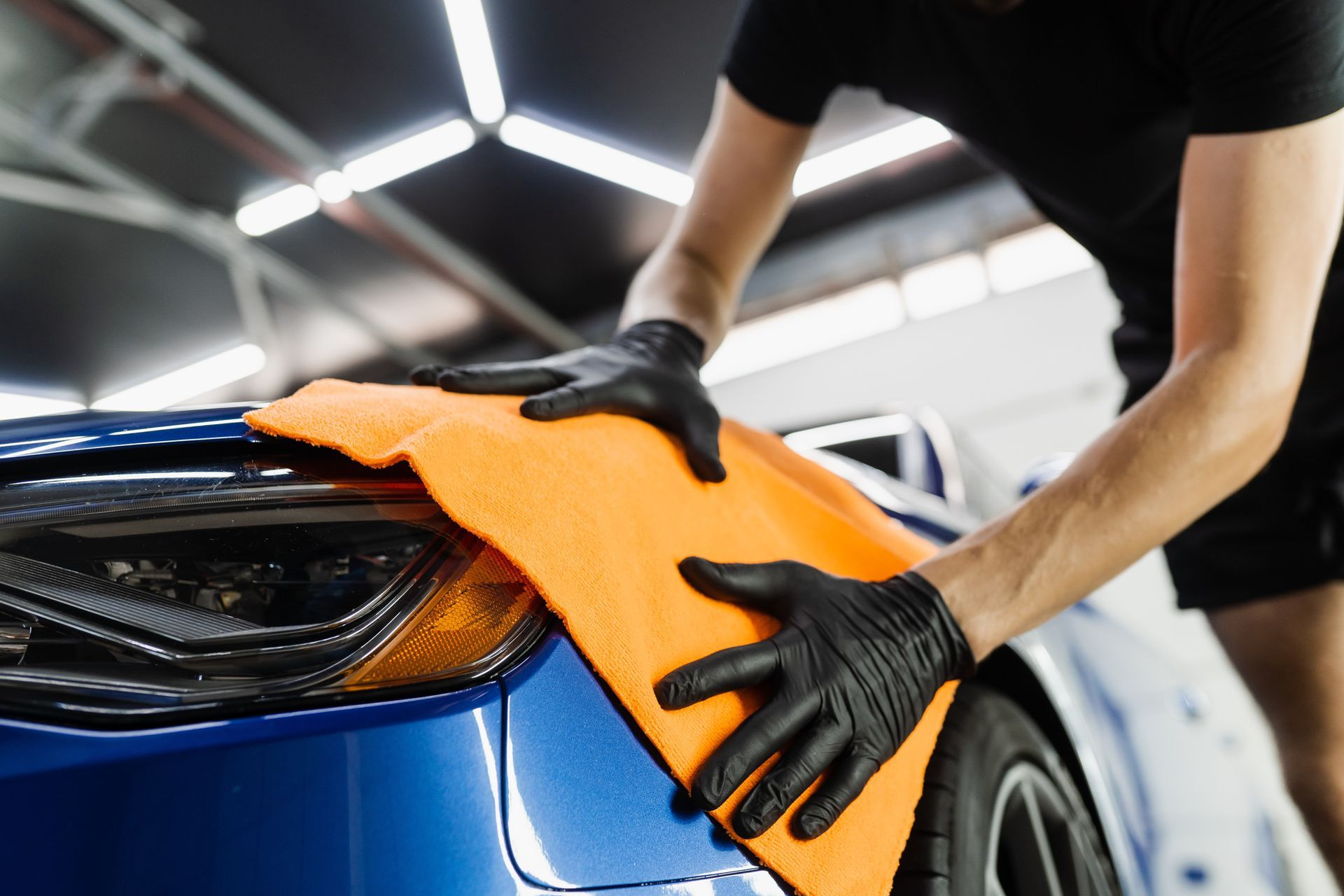 Person wearing black gloves cleaning a blue car's headlight with an orange microfiber cloth.