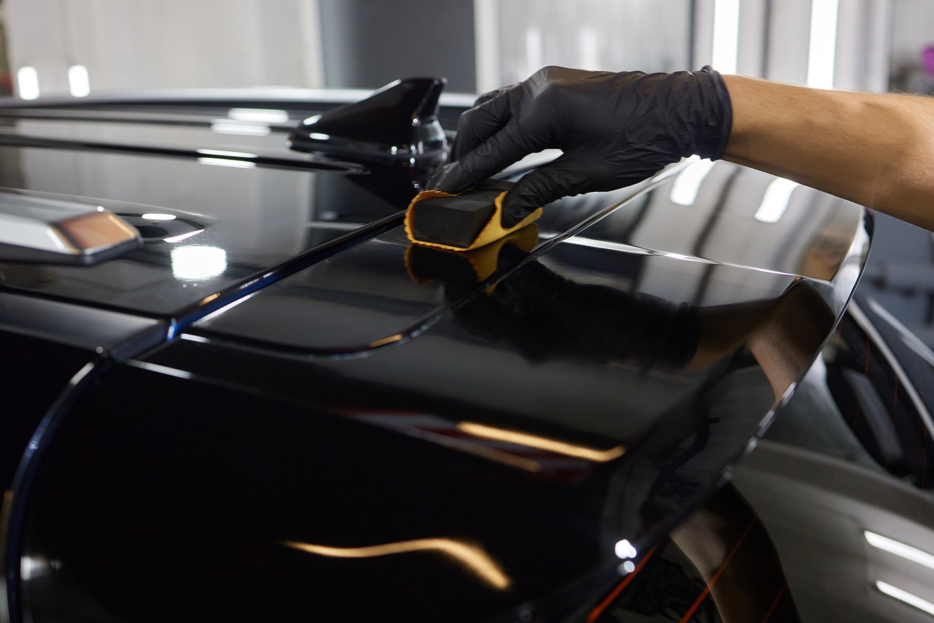 A gloved hand applies a product to a black car spoiler using a gold applicator.