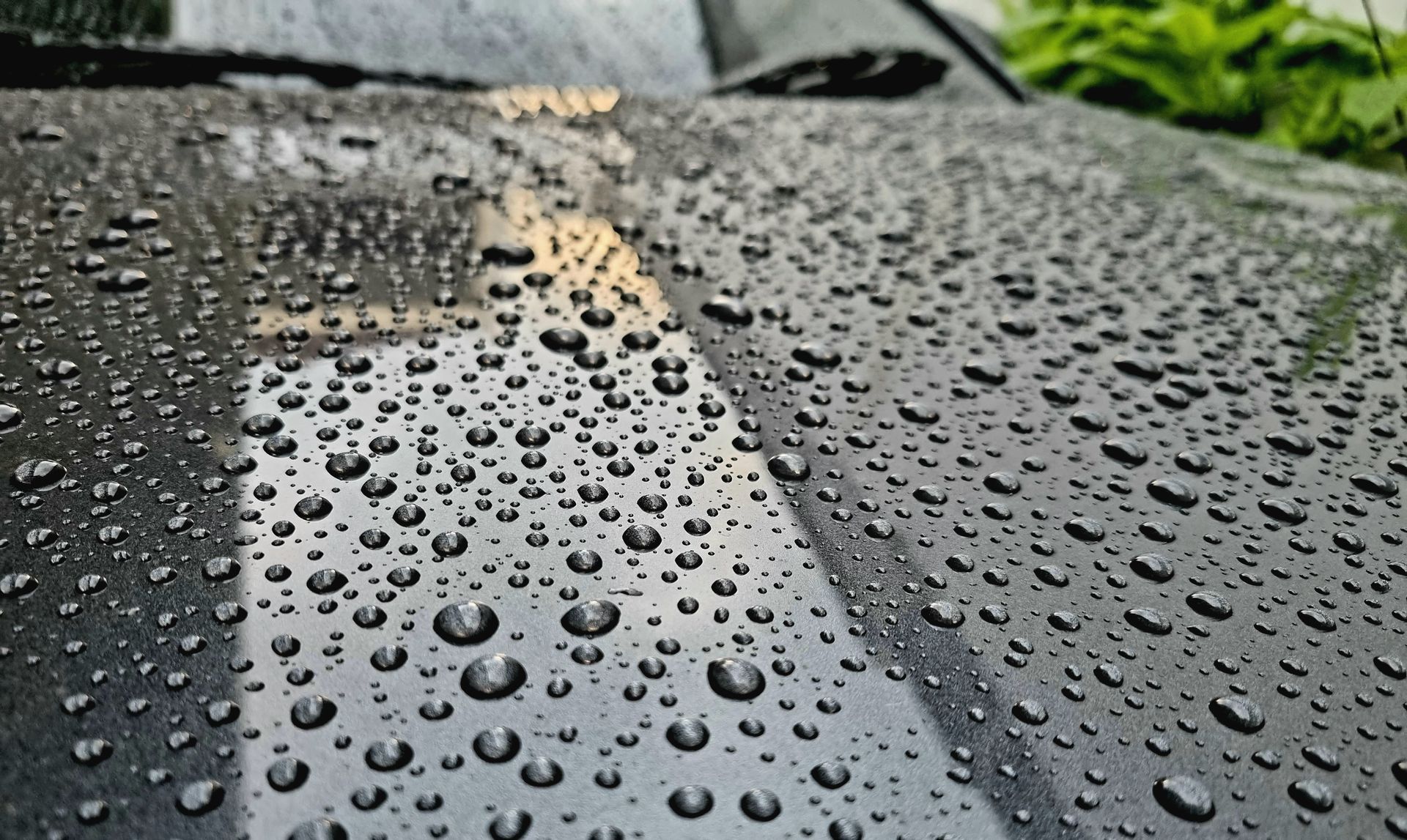 Close-up of a dark car hood covered in numerous water droplets, reflecting light.