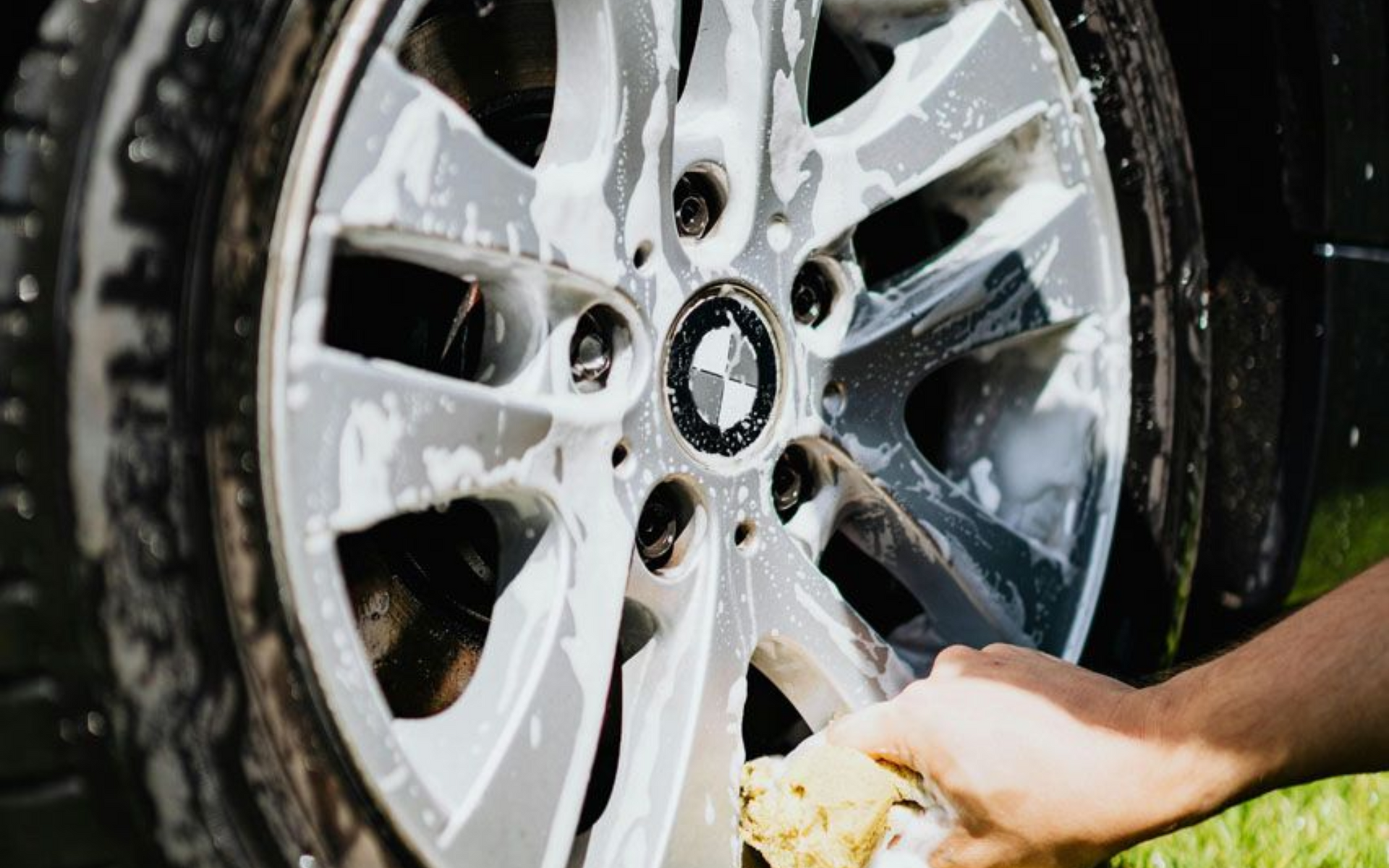 Hand washing a car's silver alloy wheel with a soapy sponge.